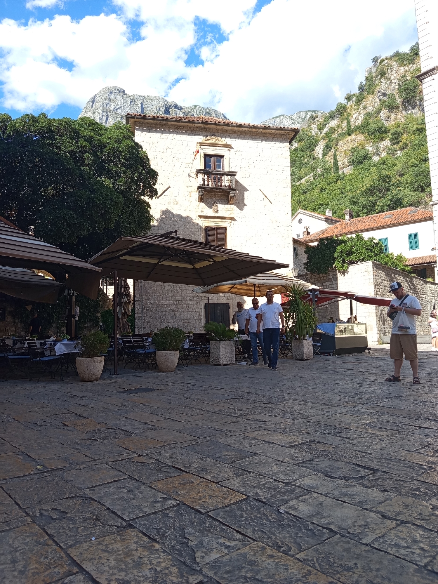 The image captures a sun-drenched European town square, likely in Kotor, Montenegro, given the mountainous backdrop. The paving stones underfoot are worn and uneven, suggesting a historic setting.
In the foreground, a man stands looking at his phone, dressed casually in a t-shirt and shorts, with sandals on his feet. Behind him, a group of three men walk across the square. The middle man, in a white t-shirt, is walking with his hand on the shoulder of the man to his left. They are all wearing casual attire.
To the left, outdoor seating areas for a cafe or restaurant are visible under large brown umbrellas. Tables are set with white tablecloths and blue chairs. Potted plants add greenery to the scene. Further back, a multi-story building with a stone facade and a balcony with an ornate railing stands prominently. The architecture suggests an older European style. Beyond this, other buildings with terracotta roofs and hints of greenery are visible, and in the far background, a majestic, rugged mountain range rises under a bright blue sky dotted with fluffy white clouds. The time of day appears to be late morning or early afternoon, judging by the strong sunlight and shadows. The overall atmosphere is relaxed and leisurely, with people enjoying the pleasant weather and the ambiance of the historic town.
