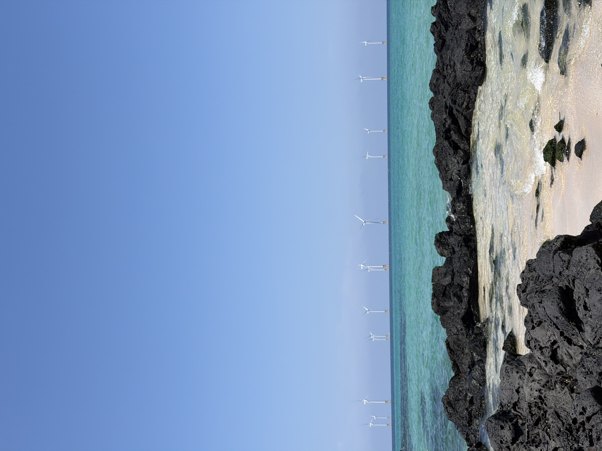 The image captures a scenic coastal view of Jeju, South Korea, under a clear blue sky. In the foreground, rough, dark volcanic rocks line the shore, with gentle waves lapping at a sandy beach. Beyond the rocks, the turquoise ocean stretches out, dotted with a line of wind turbines standing offshore. The turbines are evenly spaced and appear to be in operation, with their blades visible against the horizon. The scene conveys a sense of natural beauty and renewable energy infrastructure coexisting.