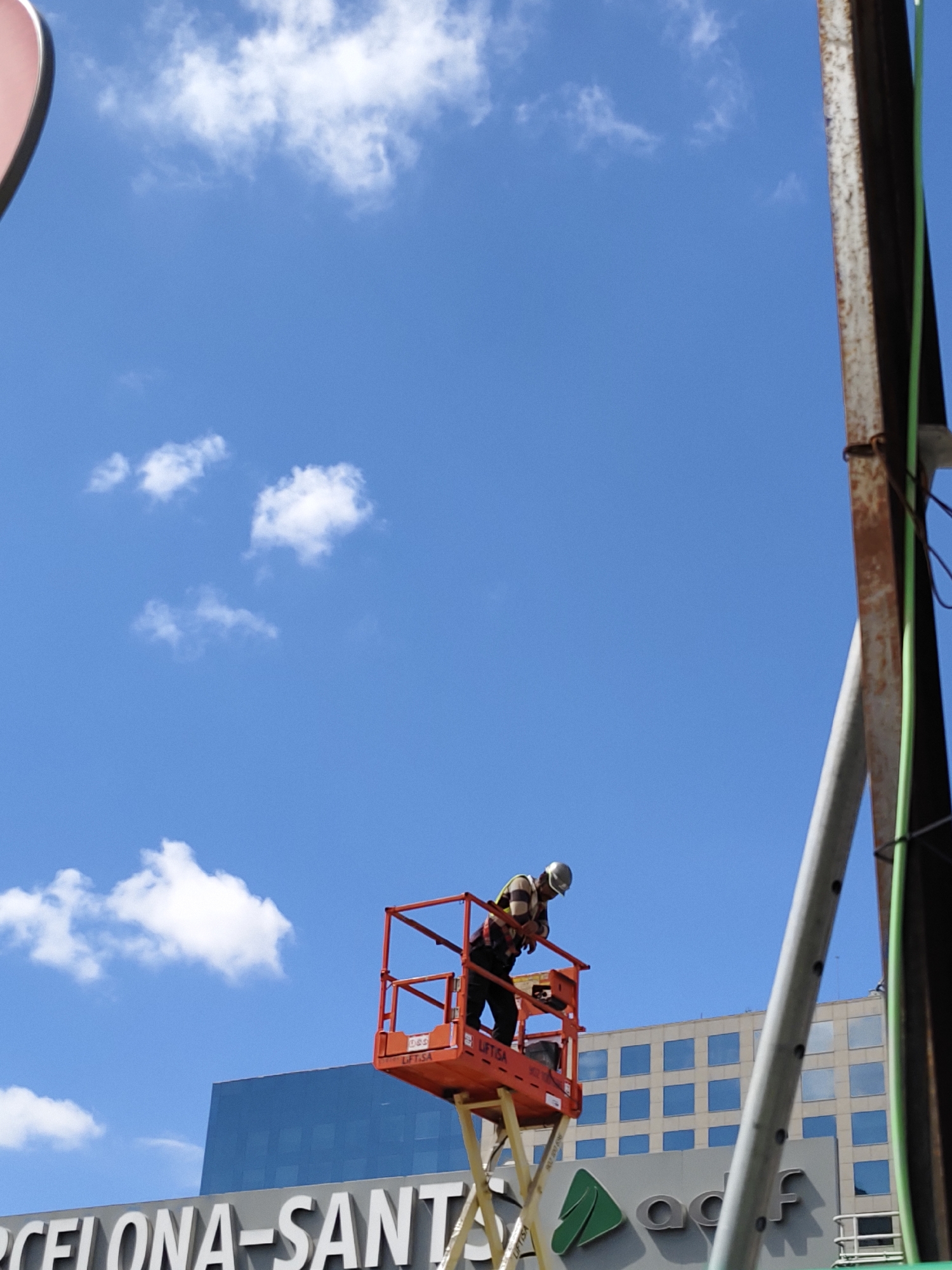 The image shows two workers in a cherry picker (aerial work platform) against a bright blue sky with scattered white clouds. The workers are wearing hard hats. The cherry picker is positioned in front of a building that has large letters spelling out "BARCELONA-SANTA" and a logo that includes green elements and the letters "adf". This suggests the location is likely near the Barcelona-Sants railway station in Barcelona, Spain. Other buildings are also visible in the background. There is also a rusty beam to the right of the cherry picker.