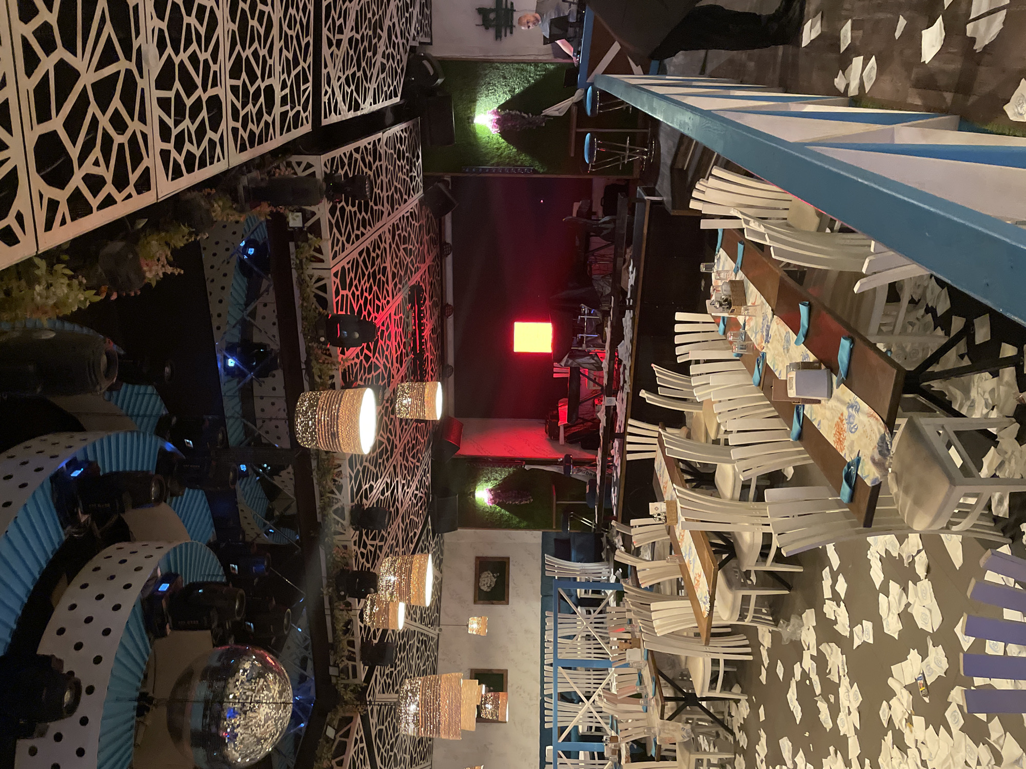 The image depicts the interior of a restaurant or bar. The location appears to be in Plovdiv, Bulgaria, based on architectural and design elements common in the region. The setting includes an outdoor or partially enclosed space with tables and chairs arranged for dining. Tables are set with blue table runners, glassware, and other dining essentials. Chairs are mostly white and wooden.
The environment shows a mix of decorative features, including light fixtures like disco balls and pendant lights. There is a stage area with lighting and audio equipment, suggesting the venue hosts live performances. A distinctive feature includes an open trellis-like structure overhead. The floor shows debris, like discarded napkins.
There are few visible people in the shot, with one individual wearing a hat partially visible.