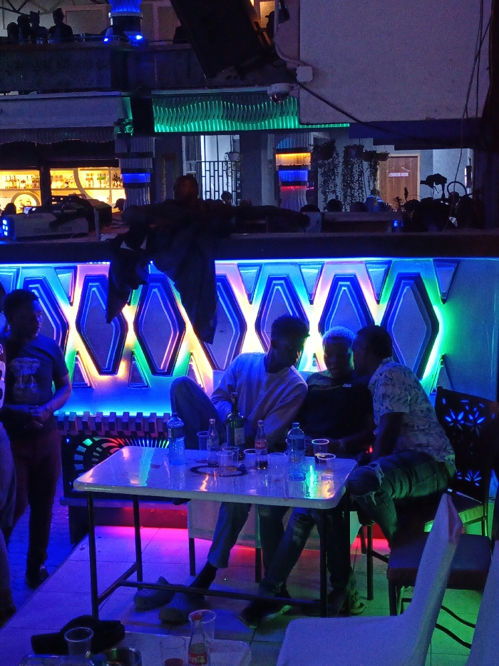 Inside a dimly lit club, four young men gather around a white table strewn with bottles and cups. The table is lit by the vibrant neon lights emanating from the bar behind them, a dazzling array of blue, green, and pink hues. A bar stocked with liquor bottles is visible in the background. Other patrons are present in the foreground and background, adding to the bustling atmosphere.