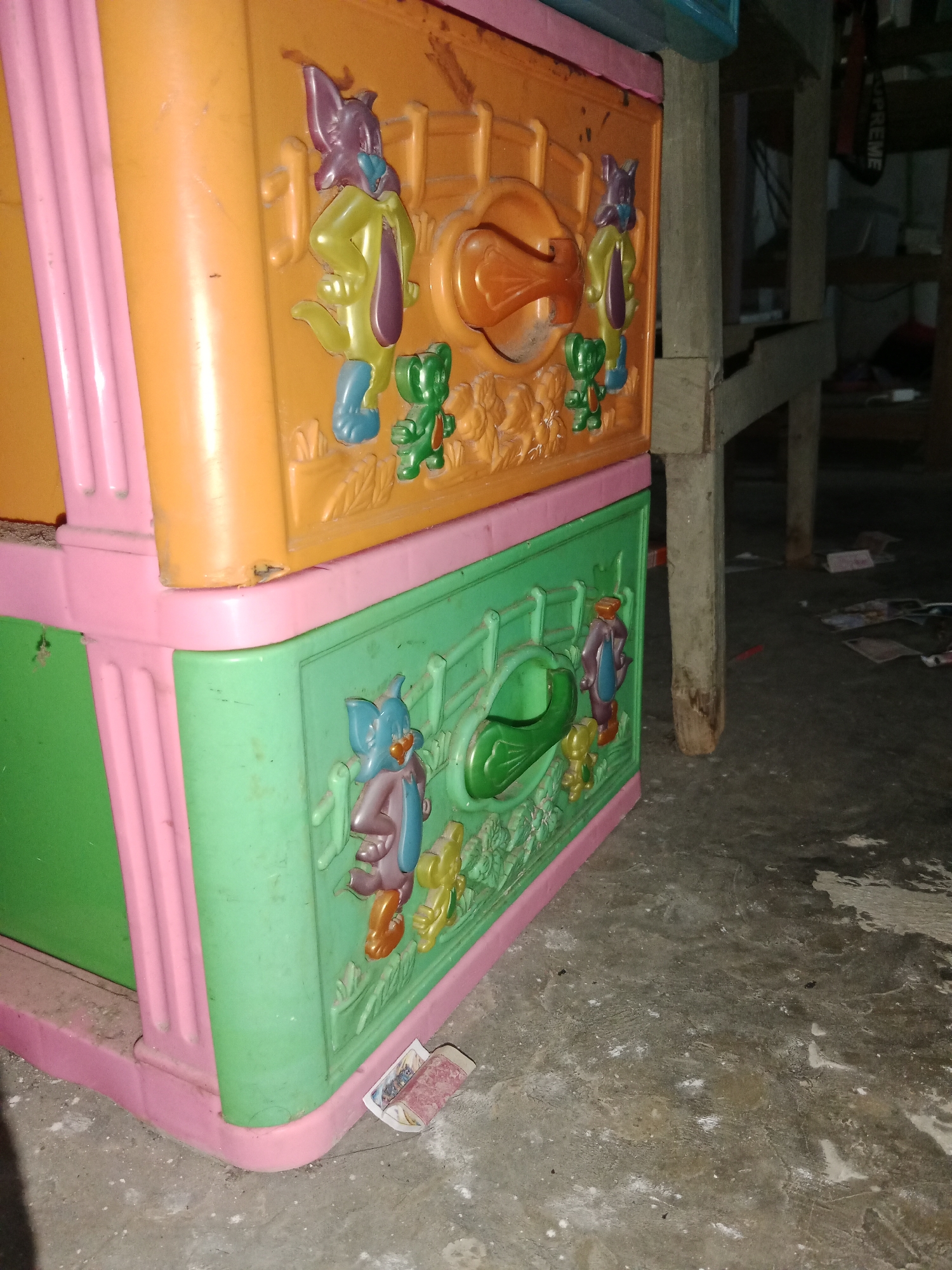 The image shows a close-up view of a pink and orange plastic drawer unit. The front of the drawers features embossed cartoon animals, including what appears to be a purple rabbit-like creature, a yellow elephant, and a green bear-like creature. The drawer unit is positioned on a concrete floor. In the background, a wooden structure and debris are visible, suggesting a cluttered or storage area. The lighting is dim, and some dust or debris is present on the floor and the plastic unit. There is no indication of any specific location in Ringinrejo, Indonesia, in the image itself.