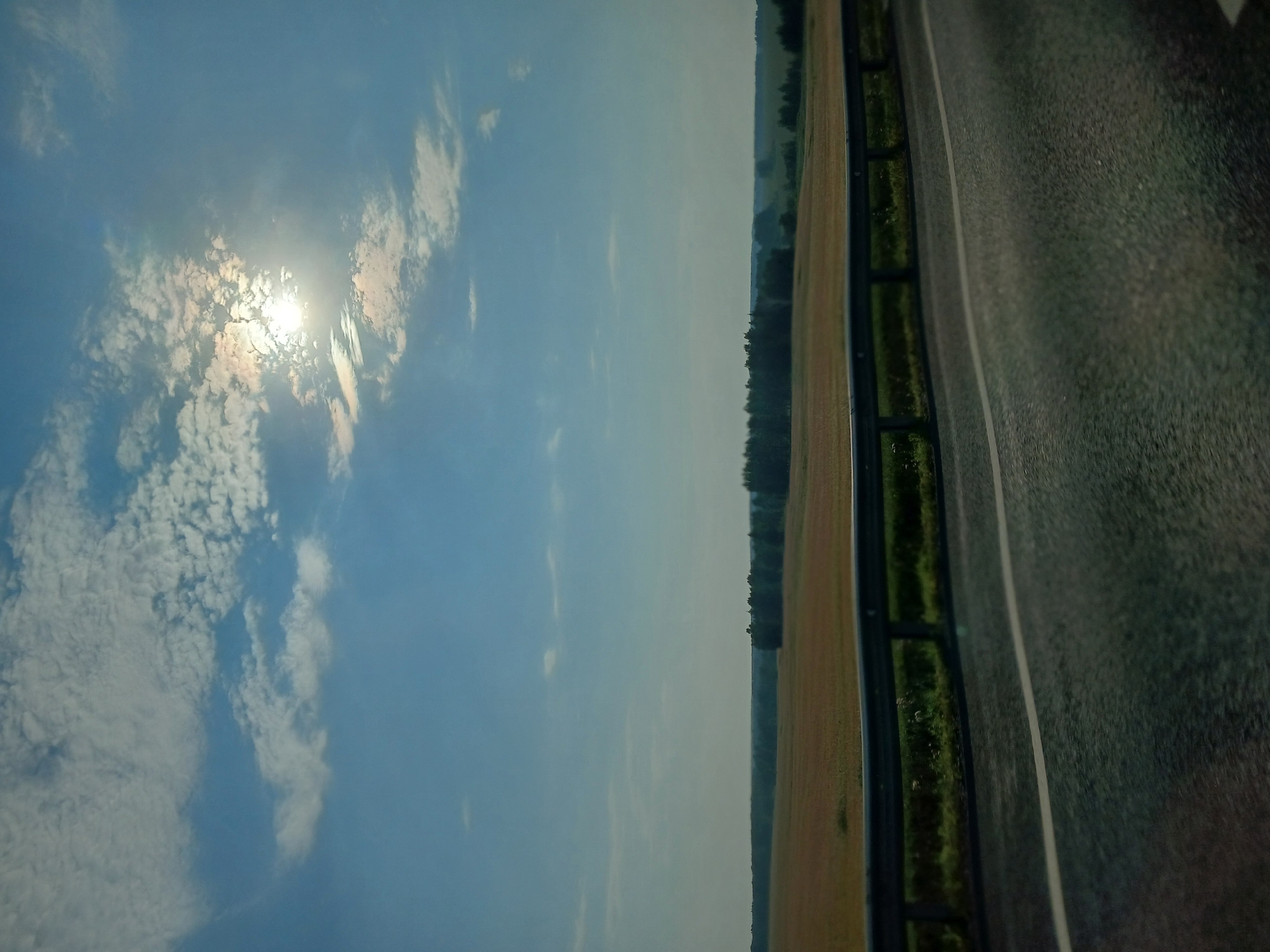 This is a landscape photograph taken from a moving vehicle on a sunny day, likely on the outskirts of Ishteryakovo, Russia. The foreground shows a portion of an asphalt road with a white lane marker. A metal guardrail separates the road from a grassy shoulder and a field. The field, appearing to be a crop of wheat or similar grain, is golden brown and stretches towards a distant line of trees forming a dark green horizon. Above the trees, the sky is a clear blue with scattered white, wispy clouds. The sun is visible in the upper right quadrant of the image, partially obscured by clouds, casting a bright glare. The scene depicts a rural, open landscape typical of the region.