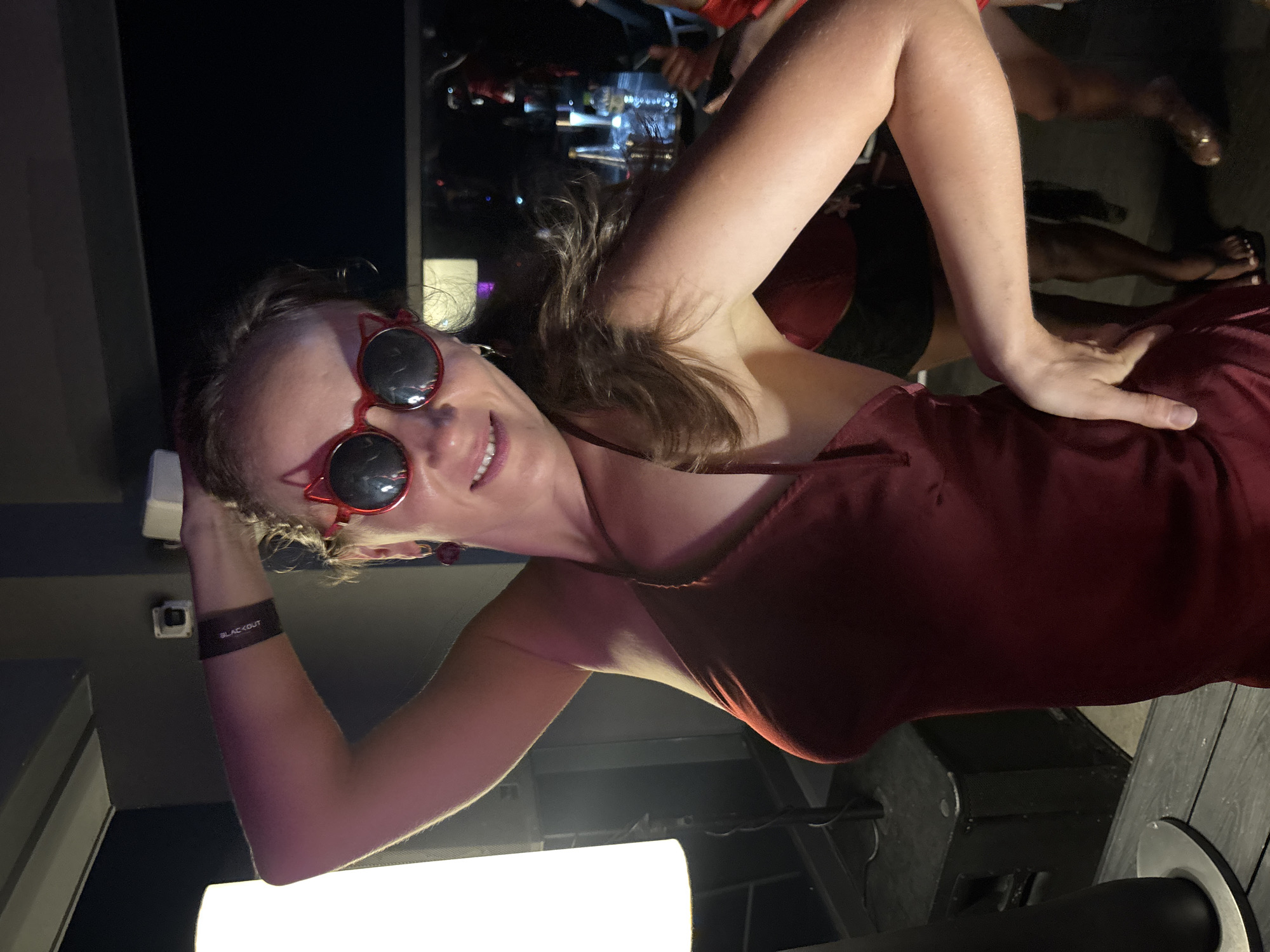 A woman in a red satin dress and cat-eye sunglasses poses with her arm behind her head. She is wearing a black wristband that reads "BLACKOUT." The background appears to be a dimly lit bar or club with other people and tables visible. The scene is set in Da Nang, Vietnam.