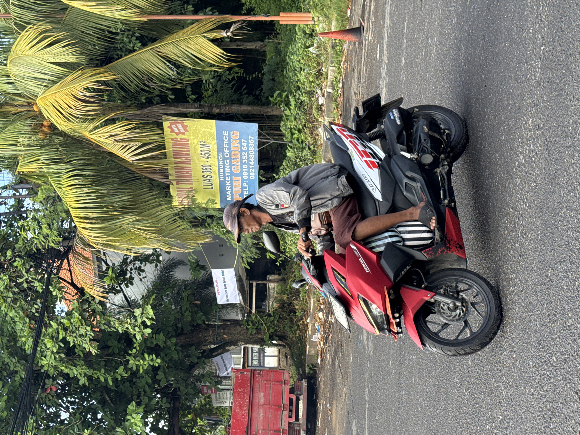 A man with a cap is riding a red Honda motorcycle on a road in Kuta Selatan, Indonesia. He is wearing sandals and striped pants. In the background, there are trees, a sign with information about Puri Gading marketing office, and a red truck. An orange traffic cone is visible on the roadside.