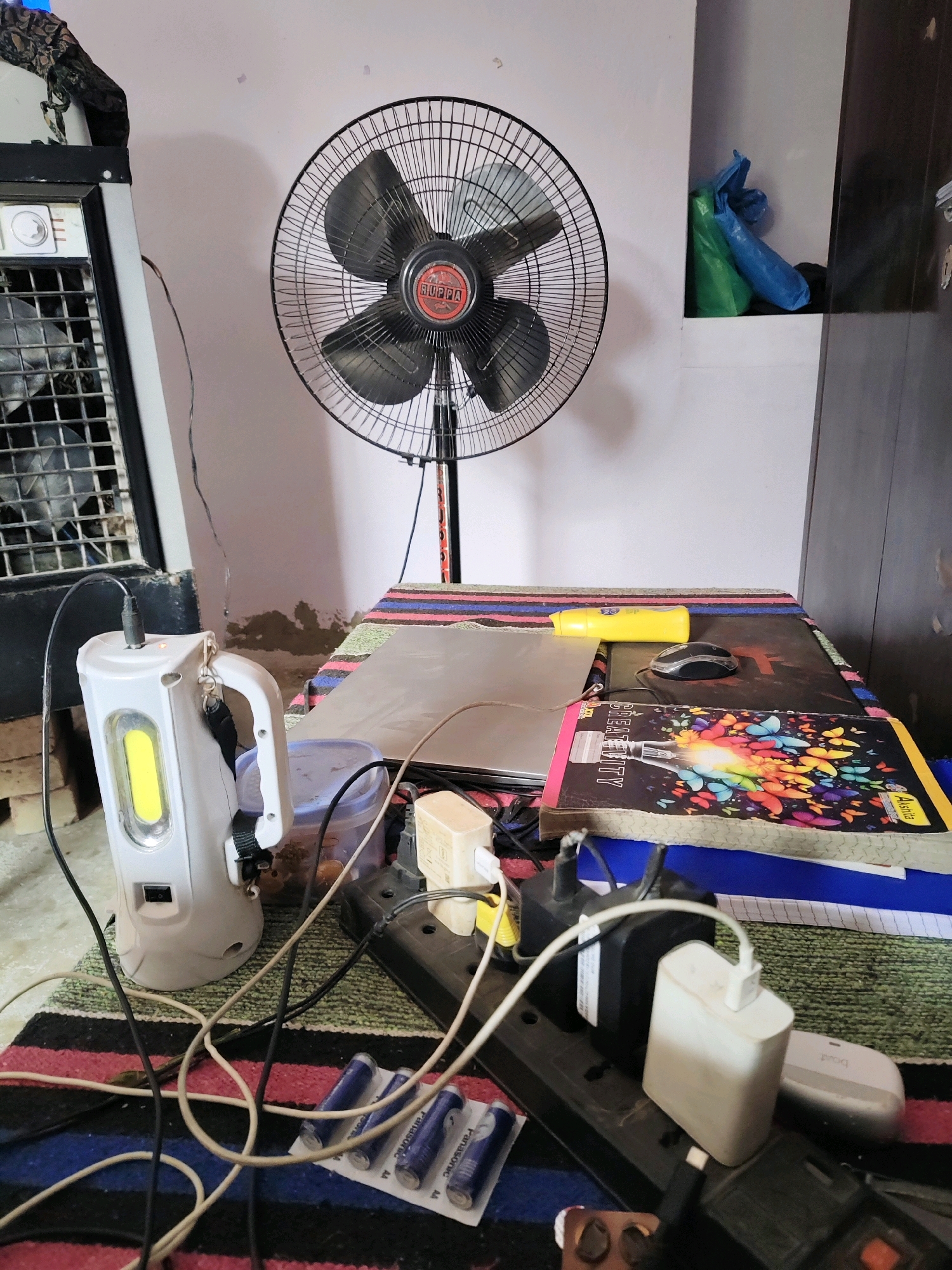 In Renwal, India, a cluttered tabletop is illuminated by a bright LED light from a white portable lamp. A black oscillating fan stands behind the table, its blades partially visible. Several electrical items are connected to a power strip, including a laptop, a computer mouse, and multiple charging adapters. A colorful notebook with a butterfly design is open on the table, alongside a yellow object that appears to be a marker or pen. A plastic container, possibly for food storage, is also present. The scene is set against a background of a light-colored wall and a portion of a portable air cooler on the left. The overall impression is one of a busy, functional workspace.