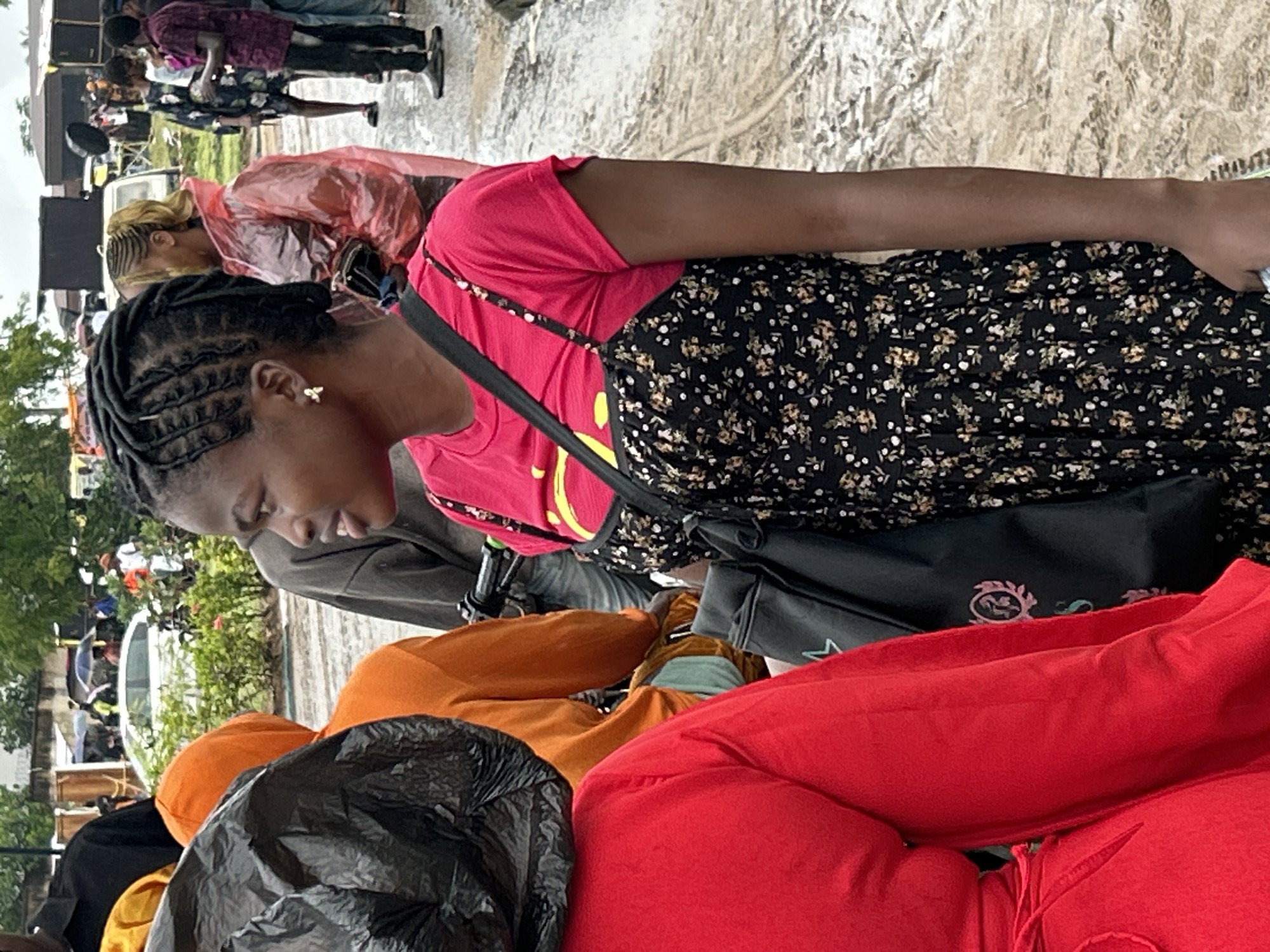 The scene is set outdoors in Ihiagwa, Nigeria, characterized by a wet, uneven dirt ground with visible puddles and reflections, indicating recent or ongoing precipitation under an overcast sky. Scattered green foliage is present throughout the midground.
In the immediate foreground, a person on the far left is partially visible, wearing a red long-sleeved garment. Directly in front of them, an individual faces away from the camera, clad in an orange hooded sweatshirt, with a black plastic bag draped over their head and shoulders.
The central subject is a young female with a dark complexion, facing left but with her head turned slightly towards the left side of the frame. She wears her dark hair styled in cornrows. Her attire consists of a bright pink short-sleeved t-shirt layered under a black spaghetti-strap dress featuring a small, light-colored floral pattern. A single gold-colored earring, designed as a bow or knot, is visible in her right ear. She carries a black shoulder bag adorned with a light blue or white emblem resembling a stylized star and leaf motif. Her right arm is visible, hanging alongside her body down to the wrist.
In the midground, behind and to the right of the central subject, a person with blonde or light brown hair tied back is partially obscured, wearing a translucent reddish-pink rain poncho and holding a small dark object in their right hand. Further right, an individual in a dark garment with a light pattern and dark pants stands, facing generally forward. To their right, a male figure in a dark patterned short-sleeved shirt and dark pants holds a small dark object in his right hand.
In the background, a white sedan is partially visible behind the orange-hooded individual and some foliage. Multiple blurry figures are scattered across the background, some utilizing umbrellas, one distinctly black umbrella visible in the distance. Dark, rectangular structures, possibly temporary buildings or shelters, are present in the far right background. A black object, possibly a tripod or camera apparatus, extends from the side of the orange-hooded person towards the central subject.