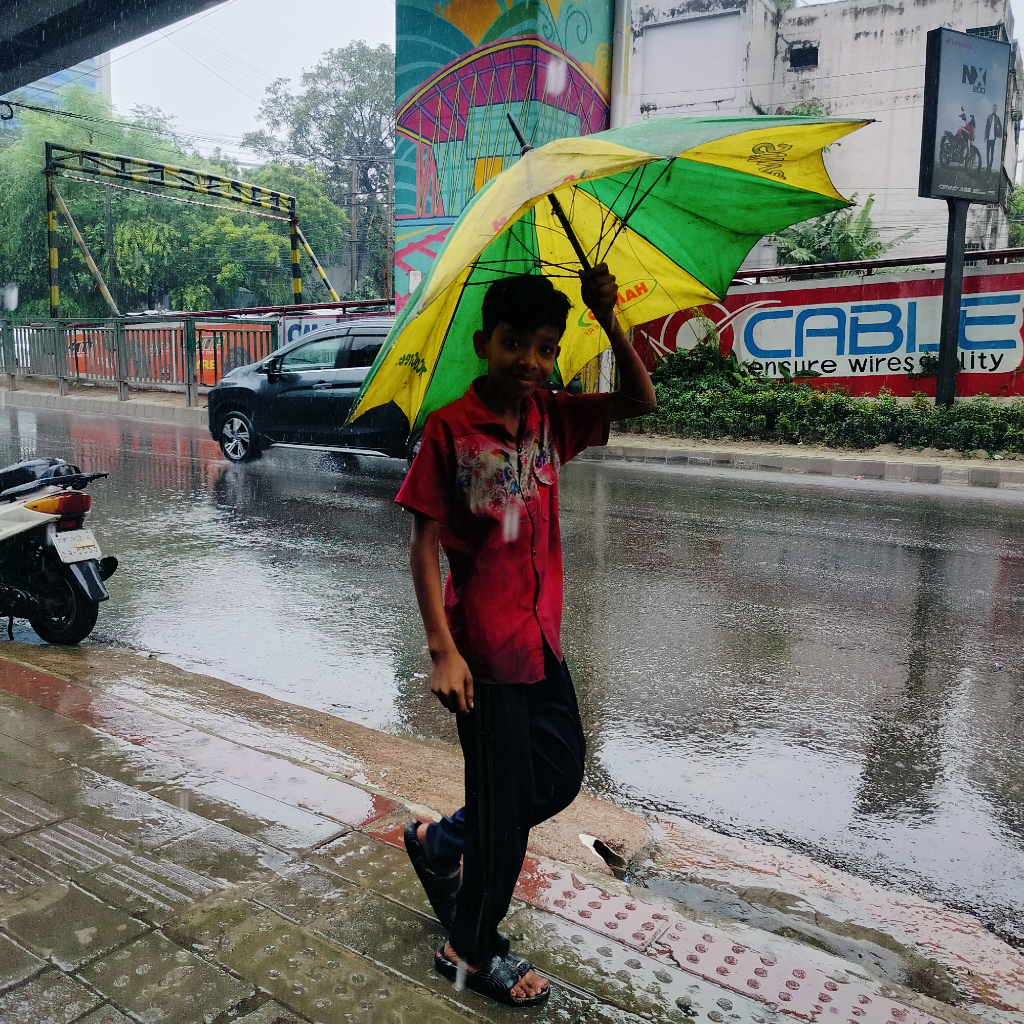 A young boy walks under a yellow and green umbrella on a rainy day in Dhaka, Bangladesh. He is wearing a red shirt with a floral pattern and black pants, and black sandals. The street is wet and reflects the surroundings. In the background, a black car is visible on the road, and a billboard with the word "CABLE" is on the right side of the image. A motorcycle is parked on the left side of the image, its rear end visible. The sky is overcast, indicating continued rainfall.