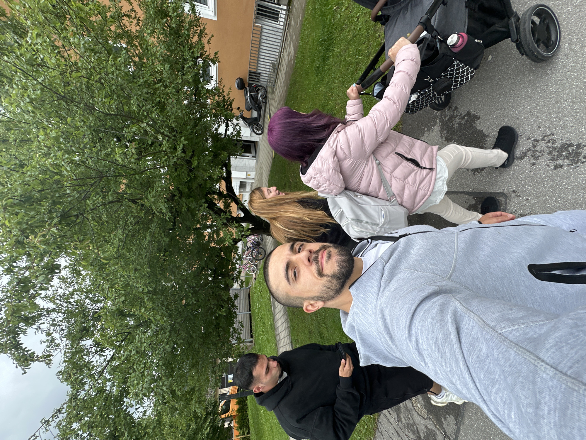 The image shows a group of people walking on a paved path in an urban area. In the foreground, a person is taking a selfie, wearing a grey hooded sweatshirt. To their left, another person in a black hooded sweatshirt is looking down at their phone. Behind them, two women are walking, one with long blonde hair and the other with purple hair. The woman with purple hair is pushing a stroller. The environment includes a grassy area, trees with green leaves, and a building with an orange facade. A parked scooter is visible in the background. The sky is overcast. There is no visual information to indicate the location is Garmisch-Partenkirchen, Germany.