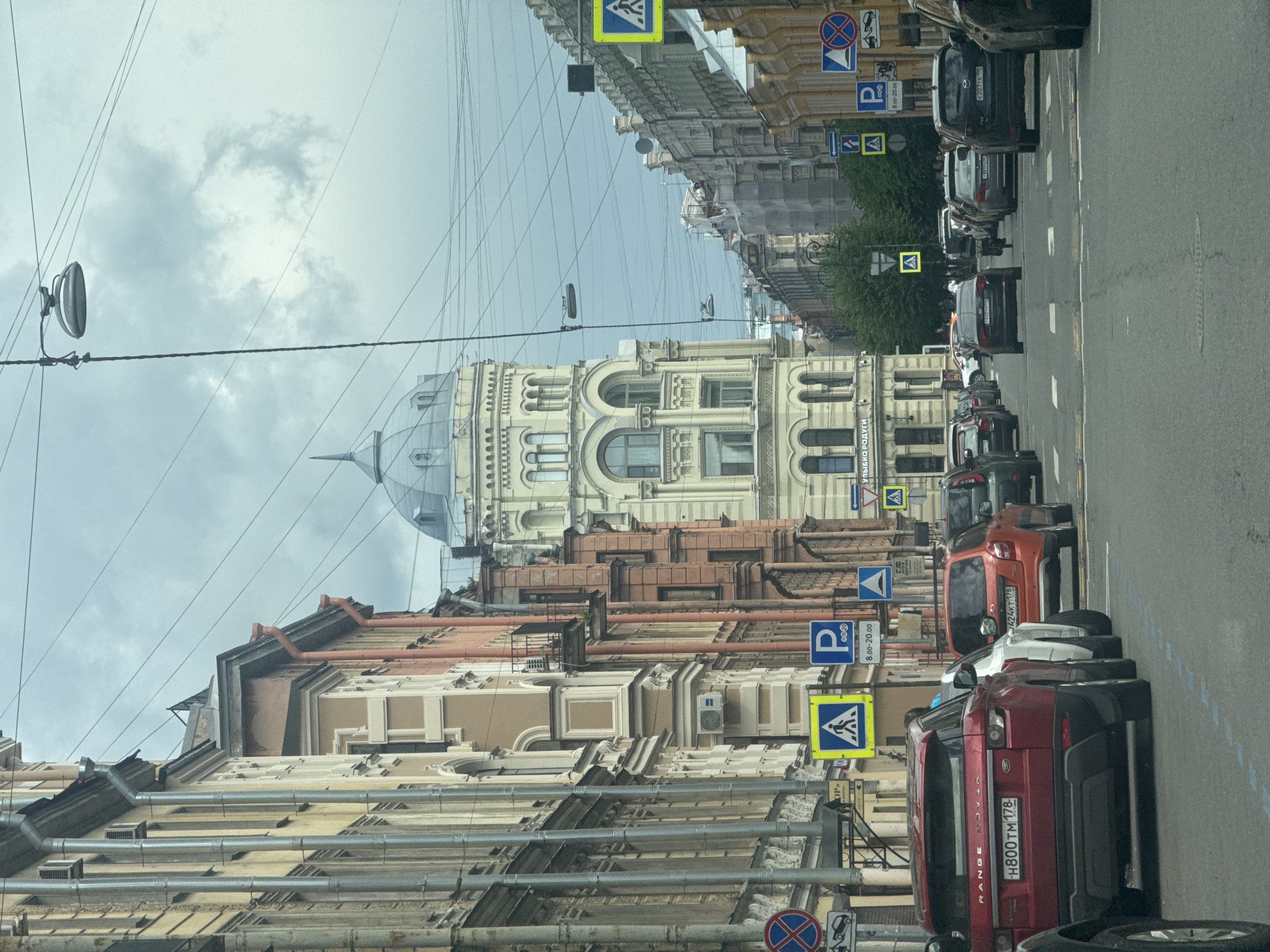The image shows a street scene in Saint Petersburg, Russia, on a cloudy day. The street is lined with multi-story buildings in an architectural style typical of the city. Several cars are driving on the asphalt road, including a red Range Rover in the foreground, an orange Lada, and other vehicles further down the street. Pedestrian crossing signs and parking signs are visible on the sidewalks. The sky is overcast with a few scattered clouds. Overhead, a complex network of electrical wires crisscrosses the sky, and a street lamp is visible. In the distance, a domed building stands out amongst the other structures. The overall impression is a view from a moving vehicle looking down a city street.