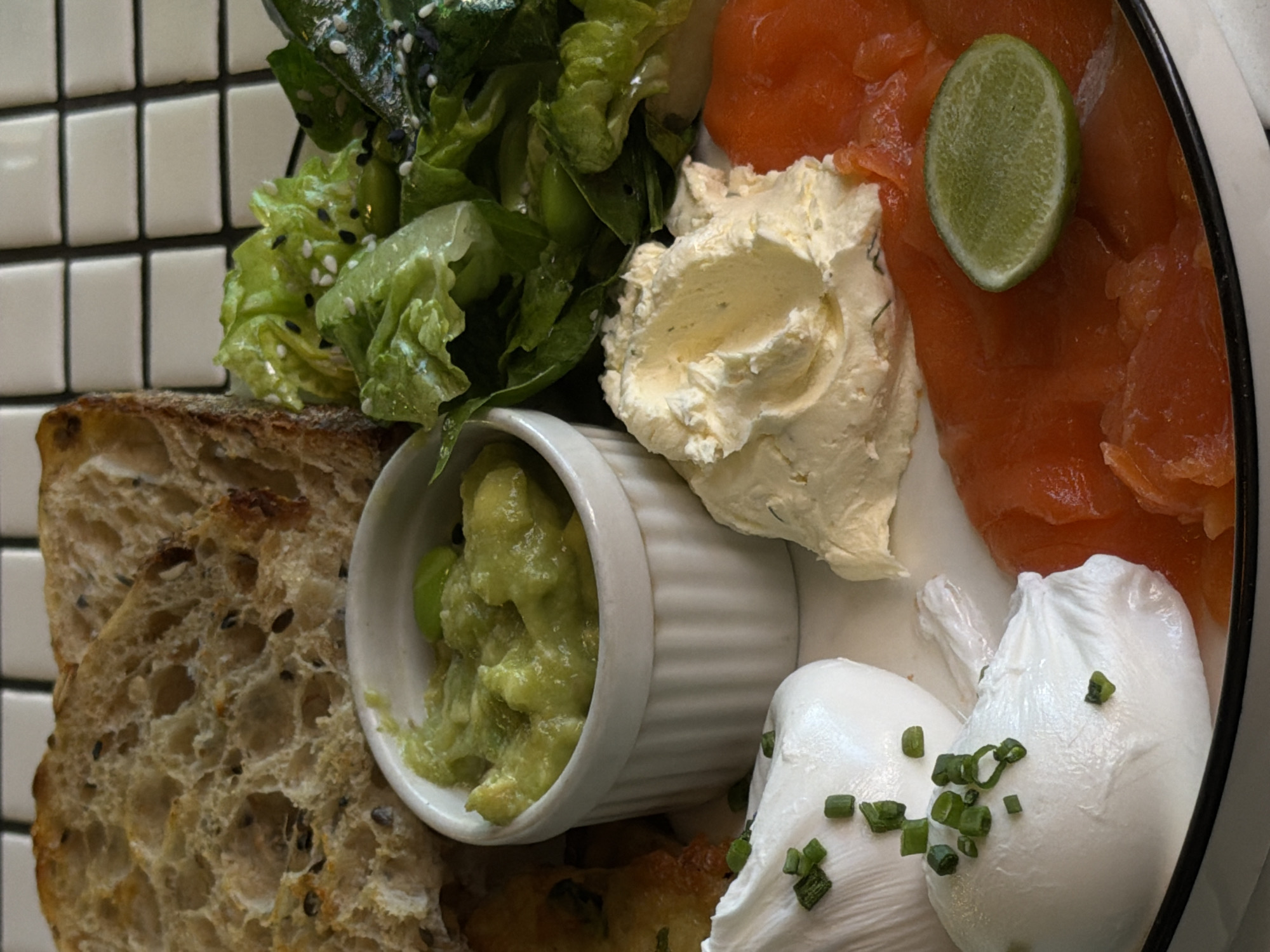 A close-up shot depicts a plate of food, featuring various components. Bread with visible seeds occupies the lower-left portion. Adjacent to it is a small white ramekin filled with a green, mashed avocado mixture. Moving to the right, there's a dollop of creamy white cheese spread, next to slices of salmon, topped with a slice of lime. The bottom-right corner shows two poached eggs garnished with chopped green herbs. A salad of green leaves and edamame beans sits at the top-left, next to the bread. The food rests on a plate with a white surface, against a background of square white tiles. The image was likely taken in North Kuta, Indonesia.