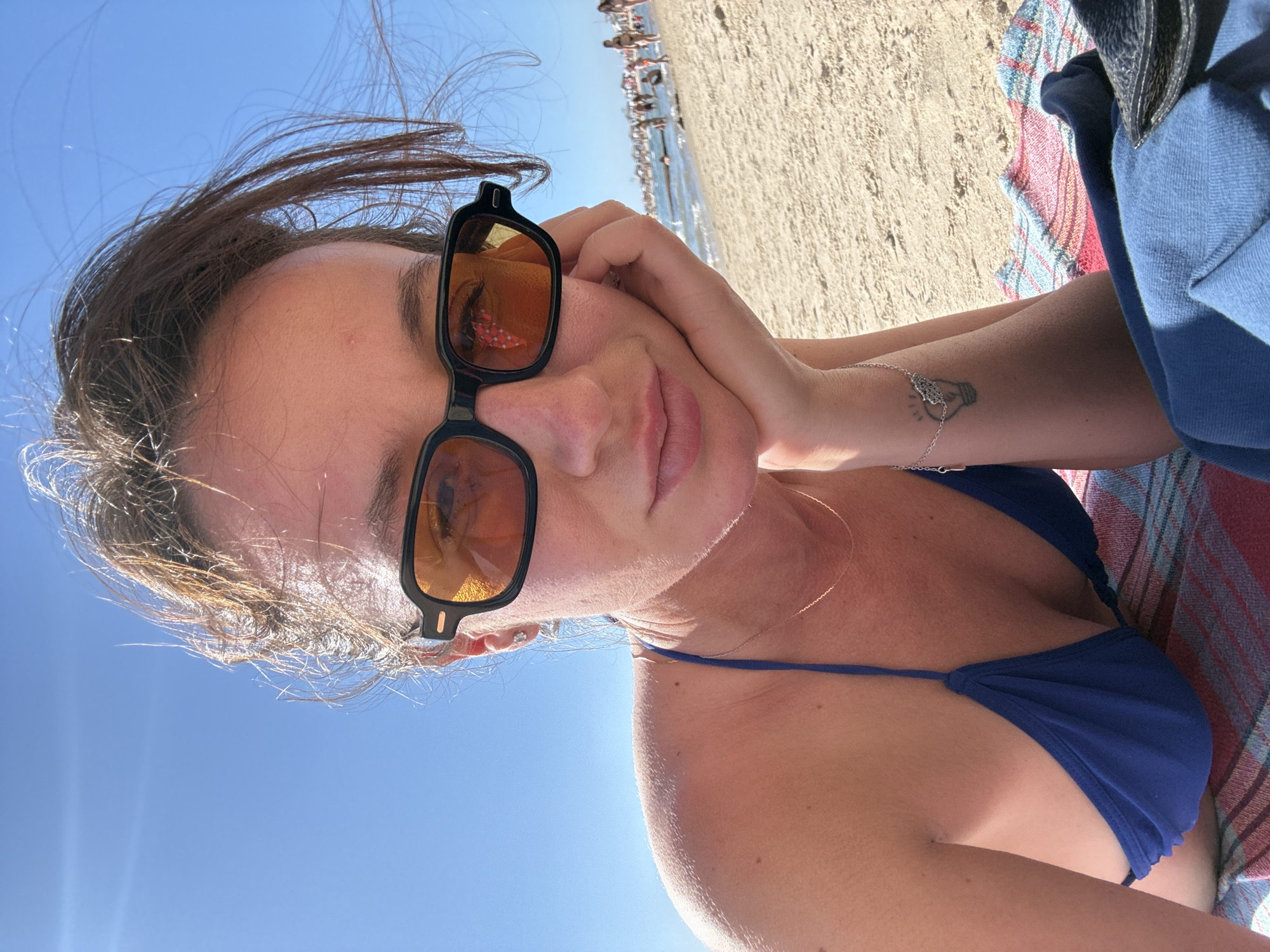A woman with dark hair, wearing a blue bikini top and orange-tinted sunglasses, lies on a striped beach towel on the sand. Her left hand rests on her cheek, and her gaze is directed towards the camera. In the background, other beachgoers are visible near the water under a clear blue sky. The scene appears to be a beach in Tel Aviv, Israel.