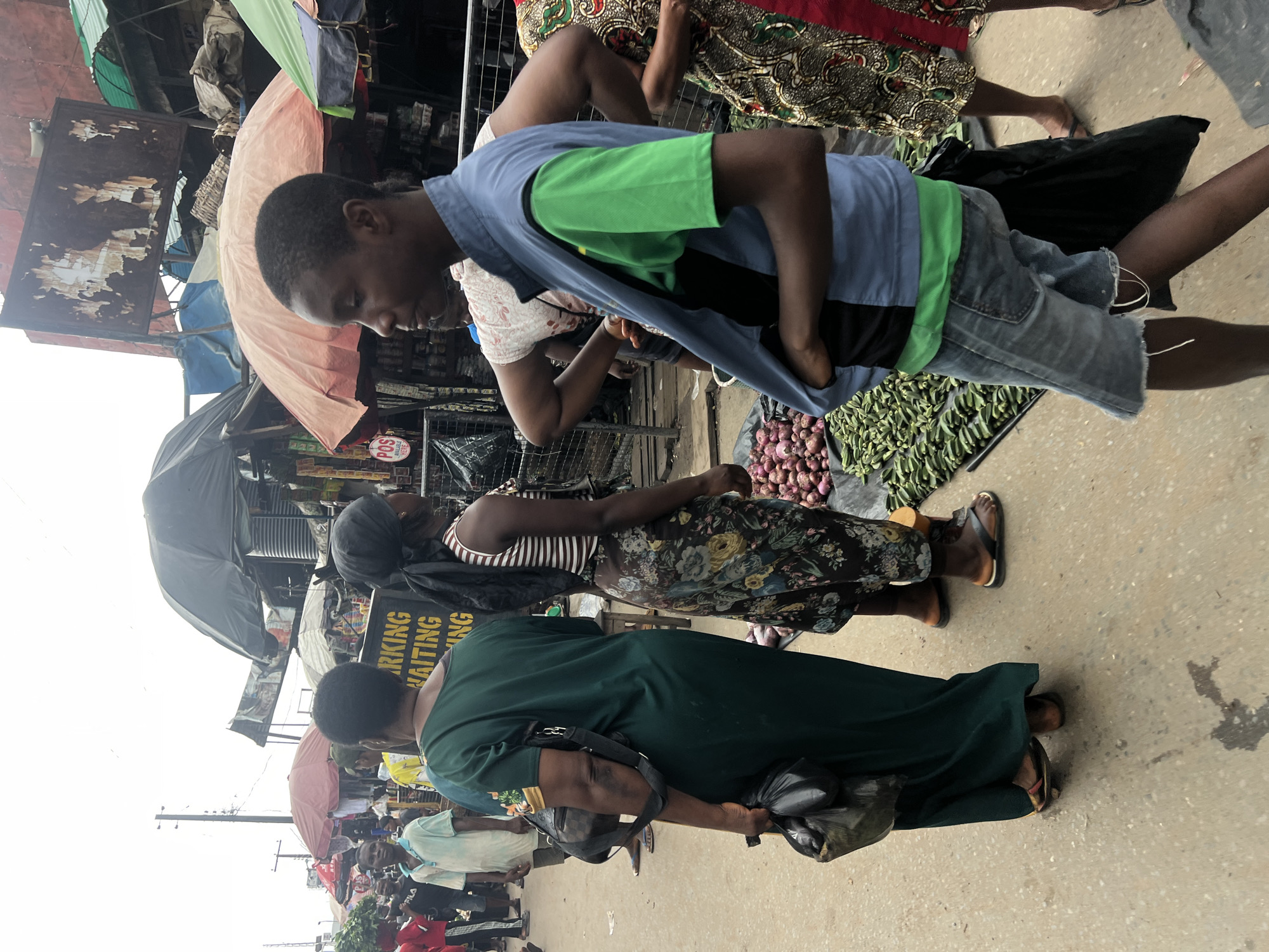 The image depicts a bustling outdoor market scene in Nkpor, Nigeria, likely along a street or thoroughfare. In the foreground, a young person in a blue vest over a green t-shirt and denim shorts is looking to the right, with their hands in their pockets. To their left, a woman in a dark green dress, carrying a black bag over her shoulder and a plastic bag in her left hand, is also facing away from the camera. Next to her, a woman in a striped top and a floral skirt stands looking forward. Behind them, various market stalls are visible, with umbrellas providing shade over displays of produce, including okra and onions. Several other people are present in the background, some appearing to be vendors and others shoppers. The ground is paved, and the overall atmosphere suggests a busy commercial area. Signs are visible in the background, including one with yellow lettering that reads "PARKING WAITING." The sky is overcast.