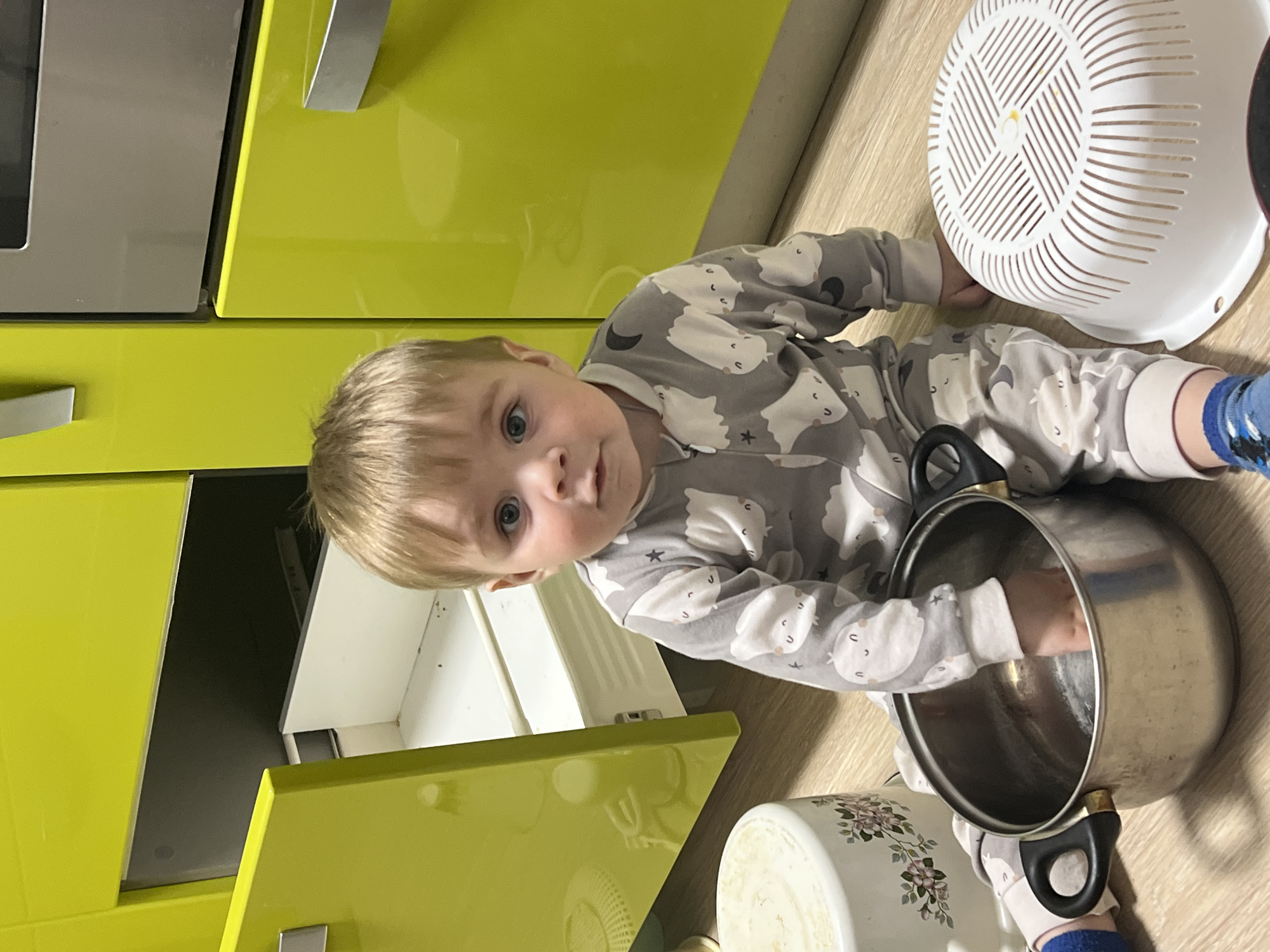 A baby, appearing to be a boy with blond hair and blue eyes, lies on a light wood-effect kitchen floor. He is dressed in a grey onesie with white cloud and moon patterns and blue socks. The child looks directly at the camera with a curious expression.
He is surrounded by kitchen items, seemingly playing. His right hand is inside a large silver metal pot with black handles. A white plastic colander with a spiral design rests near his left arm, and part of another white pot with a floral pattern is visible at the bottom of the frame.
The background features bright, glossy lime green kitchen cabinets with metal handles. One cabinet door or drawer is slightly ajar, revealing a dark interior. Part of a silver kitchen appliance, possibly an oven or microwave, is visible on the far left. The setting is an indoor kitchen, well-lit as if during the daytime, likely a home in Perm, Russia. There is no visible text in the image.