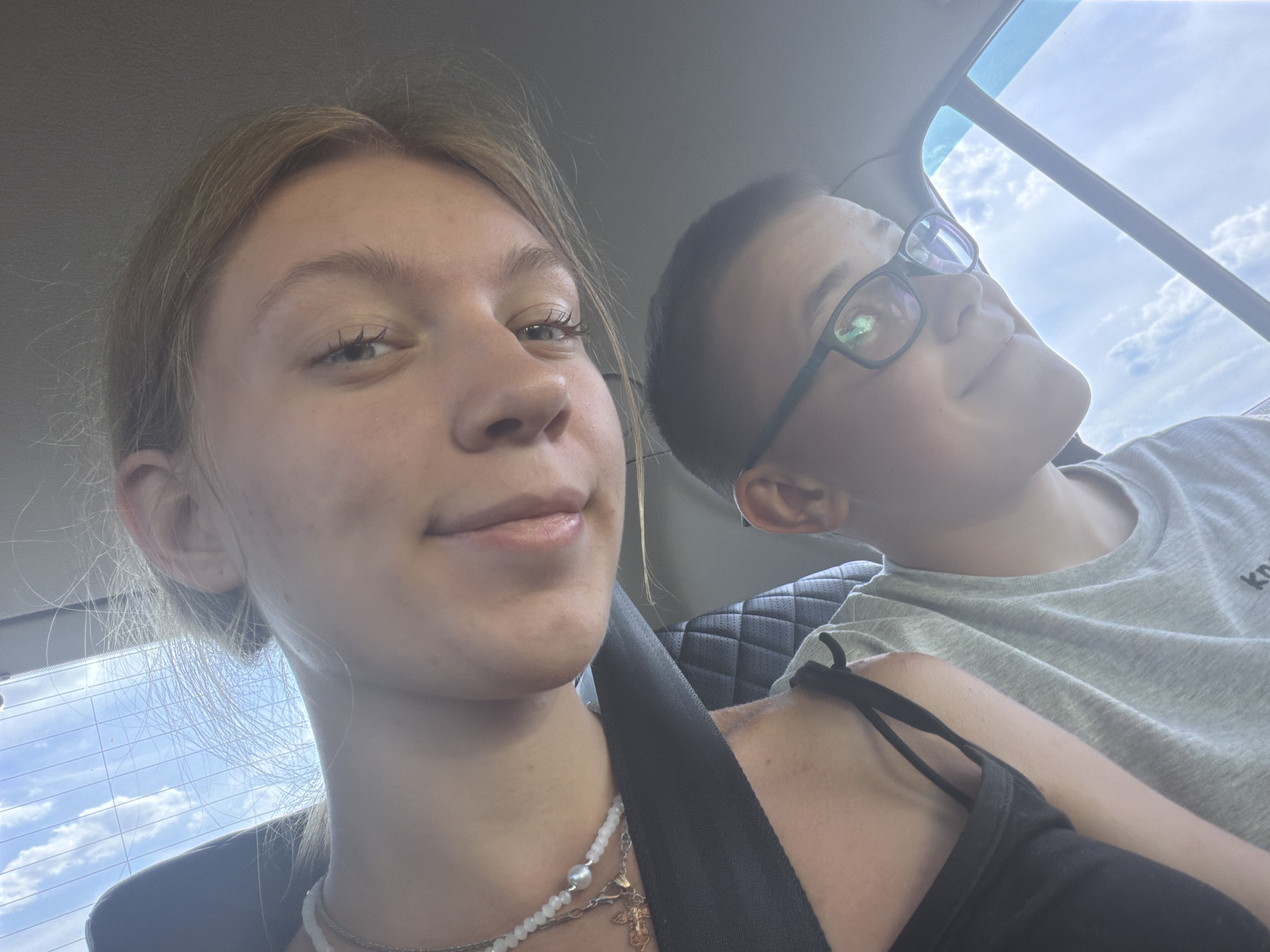 Two individuals, a female in the foreground and a male in the background, are captured in a selfie taken inside a vehicle. The female, positioned closer to the camera, has blonde hair and is wearing a black top and a pearl necklace. She is looking directly at the camera with a slight smile. The male, wearing glasses and a grey t-shirt, is seated behind her, looking upwards. The interior of the car is visible, with a black leather seat and a seatbelt. The background reveals a cloudy sky through the car window, suggesting an outdoor setting. The image was taken in Ostrovka, Russia.