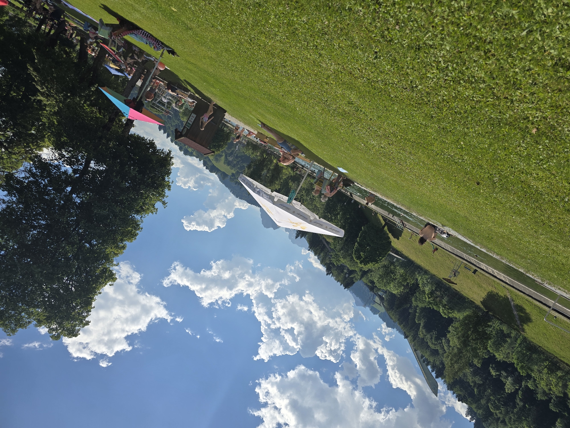 The image depicts a sunny day at an outdoor swimming area, possibly a lido or pool. The scene is filled with people enjoying recreational activities.
The main subjects are numerous individuals sunbathing, sitting, and possibly swimming. Objects visible include lounge chairs, umbrellas, a children's play structure with a slide, and a wooden building that appears to be a changing room or snack bar. The swimming area itself is a long, rectangular pool or channel. The surrounding environment is characterized by lush green grass, dense trees, and a mountainous backdrop, which, along with the specific context of Garmisch-Partenkirchen, Germany, suggests a scenic alpine location.
The activity taking place is clearly leisure and recreation, centered around a swimming facility. The time of day appears to be midday or early afternoon, indicated by the bright sunlight and clear blue sky dotted with fluffy white clouds. The weather is fair and warm, ideal for outdoor activities.
Notable details include a large, white parasol with visible text, although the text is partially obscured and difficult to read clearly. There are also hints of text on the wooden building. The overall mood conveyed is one of relaxation and enjoyment on a pleasant summer day. The composition of the image is dynamic, with the landscape tilted, giving a unique perspective.