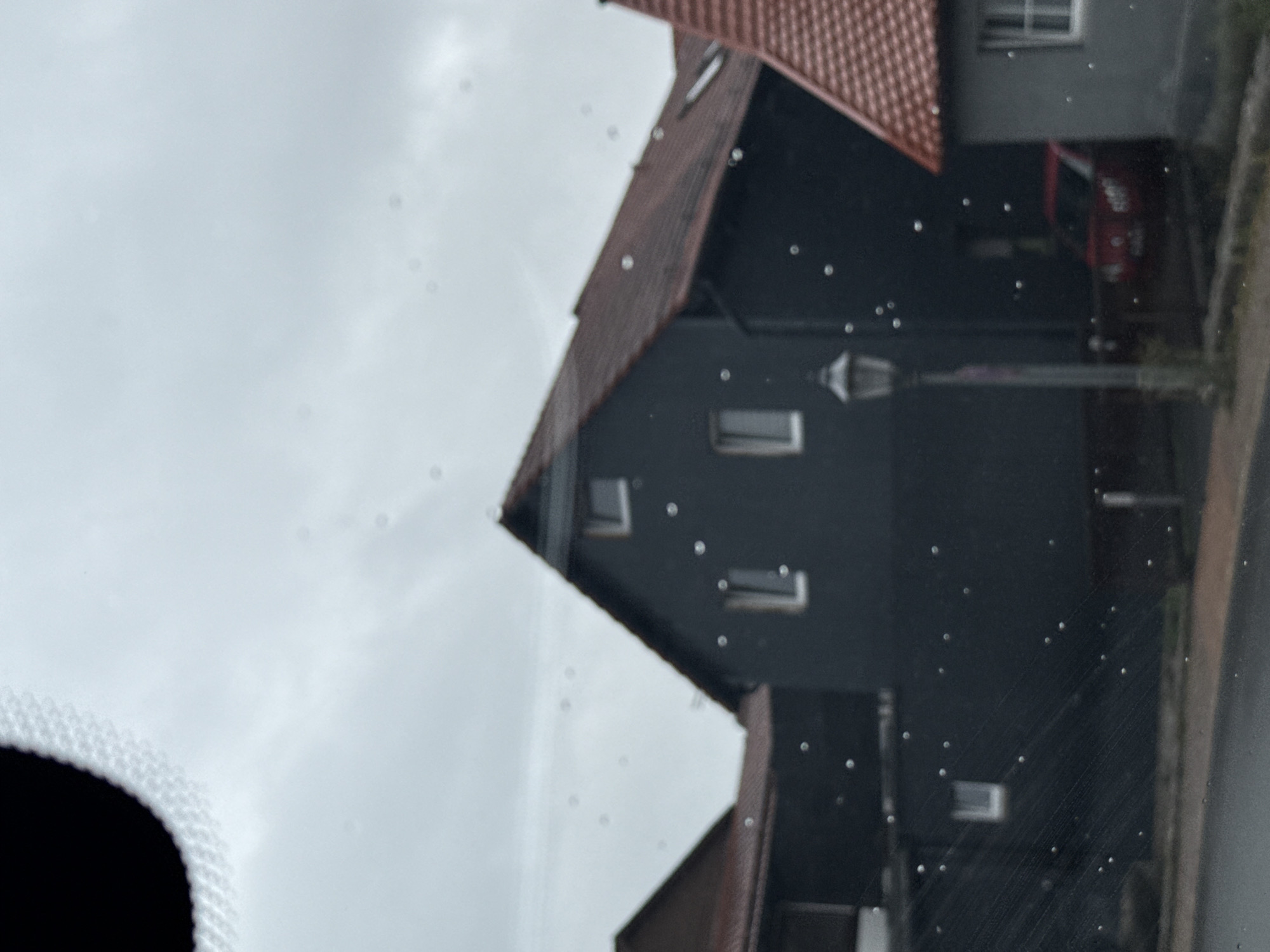 The image captures a street scene in the town of Bad Sachsa, Germany, under an overcast sky. In the foreground, the view appears to be through a window, with numerous water droplets or imperfections visible on the glass. A dark-colored building with a red-tiled roof dominates the center of the frame. This building has several windows, some of which are illuminated. To the left, a portion of another building, also with a red-tiled roof, is visible. To the right, a smaller gray building with a red-tiled roof and a white-framed window is present. A red car is parked behind a fence between the central and right buildings. A street lamp stands in front of the dark building, and a paved road or sidewalk is visible in the lower portion of the image.
