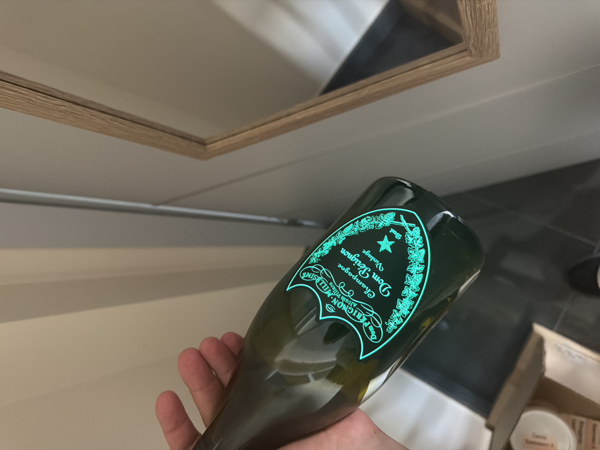 A person's hand holds a green glass bottle of Dom Pérignon champagne. The bottle has a glowing turquoise label with the brand name, "Dom Pérignon," "Champagne," and "Vintage" printed on it. A star emblem is also visible on the label. The background consists of a wall and a wooden-framed mirror. The scene is indoors, likely in a residential setting, and appears to be in Dolno Ablanovo, Bulgaria. The lighting is dim, with the bottle's label being the brightest element.