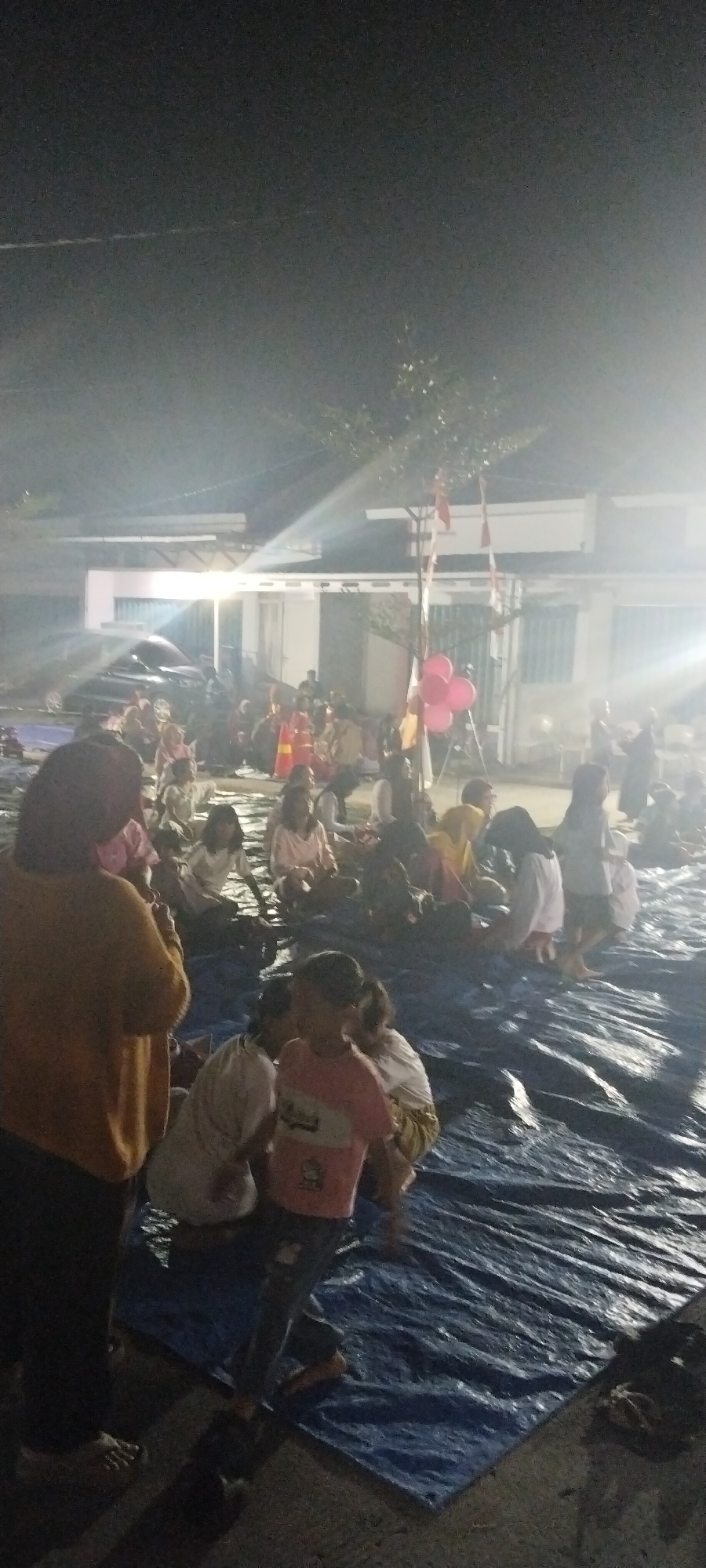 The image depicts an outdoor gathering at night in what appears to be a residential or community area in Taban, Indonesia. A group of people, predominantly children, are seated on a large blue tarp spread on the ground. The lighting suggests artificial illumination, possibly from floodlights, casting strong beams and creating glare in the upper portion of the frame. In the background, there is a building with white walls and a dark roof. Several flags, red and white, are visible on poles, along with pink balloons, indicating a festive or celebratory occasion. A dark-colored vehicle is parked near the building. The ground surface appears to be paved. The overall atmosphere is that of a communal event taking place after dark.