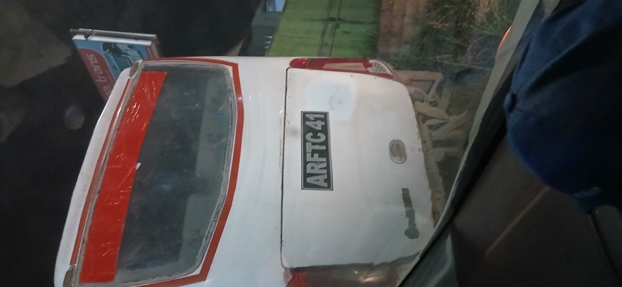 The image shows the rear of a white bus with a red stripe around the top of the rear window. A license plate with the text "ARFTC 41" is mounted centrally on the bus's rear. The bus appears to be parked or moving slowly under an overhead structure, possibly a bridge or overpass, as evidenced by the dark ceiling above. To the upper right, a billboard or sign with the text "e trans" is visible. The scene is outdoors, with some greenery and a fence-like structure visible in the background, suggesting an urban or roadside environment. The lower part of the image shows a dark interior, likely the cabin of another vehicle, with a dark blue cap in the foreground. The overall lighting suggests it is either dusk or nighttime. The bus's rear bumper and lower body are dusty. There are no people clearly visible in the image. Given the license plate format and common bus brands in the region, this scene is likely located in Samarinda, Indonesia.
