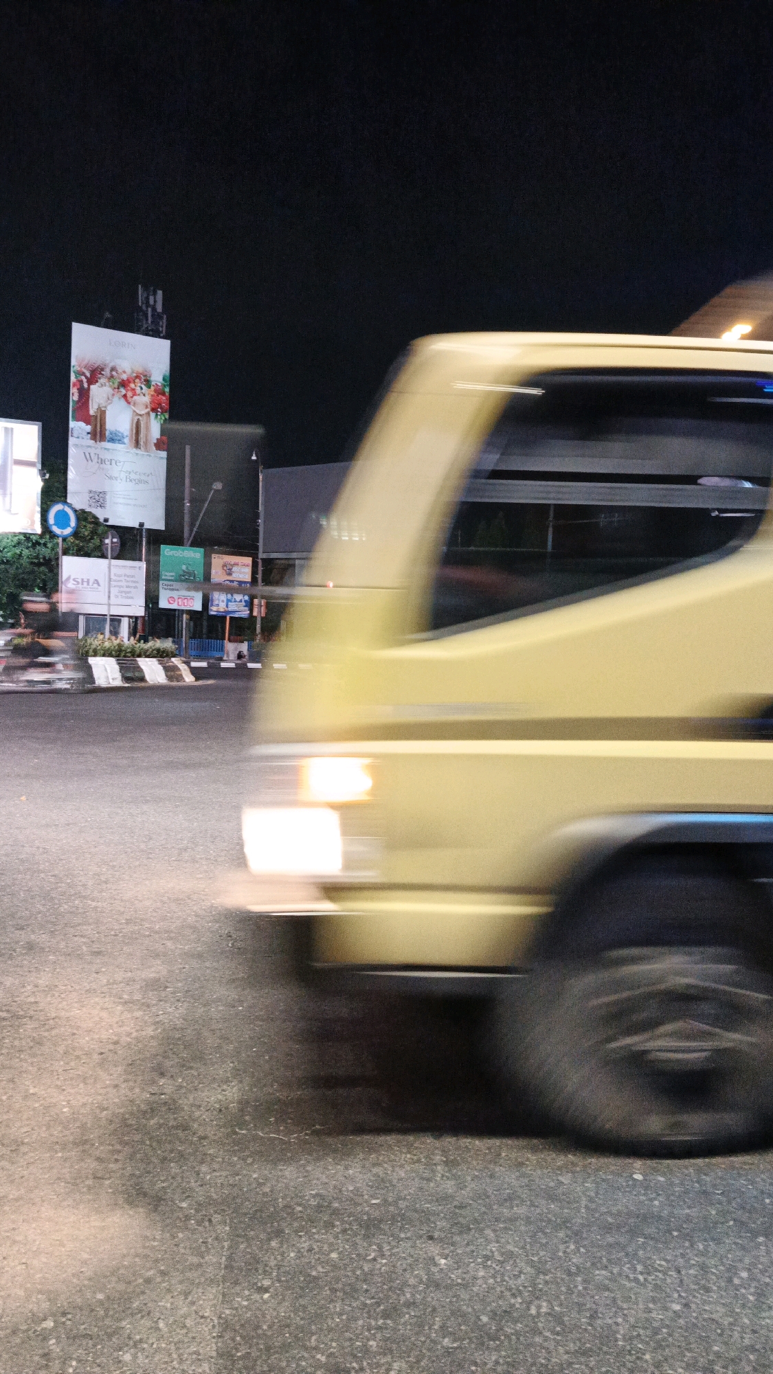 A yellow truck is captured in motion at night on a street in Surakarta, Indonesia. The image is a long exposure, resulting in motion blur for the truck and the surrounding environment. The truck's headlights are illuminated, casting a glow on the asphalt road. In the background, several billboards are visible. One billboard, positioned higher than the others, features an advertisement with text that appears to read "LORIN" at the top and a scene of a couple in traditional attire with the text "Where... Stor's Begins" below. Other smaller signs are also present, including one with "GrabBike" and another with "SHA". The sky is dark, indicating it is nighttime.