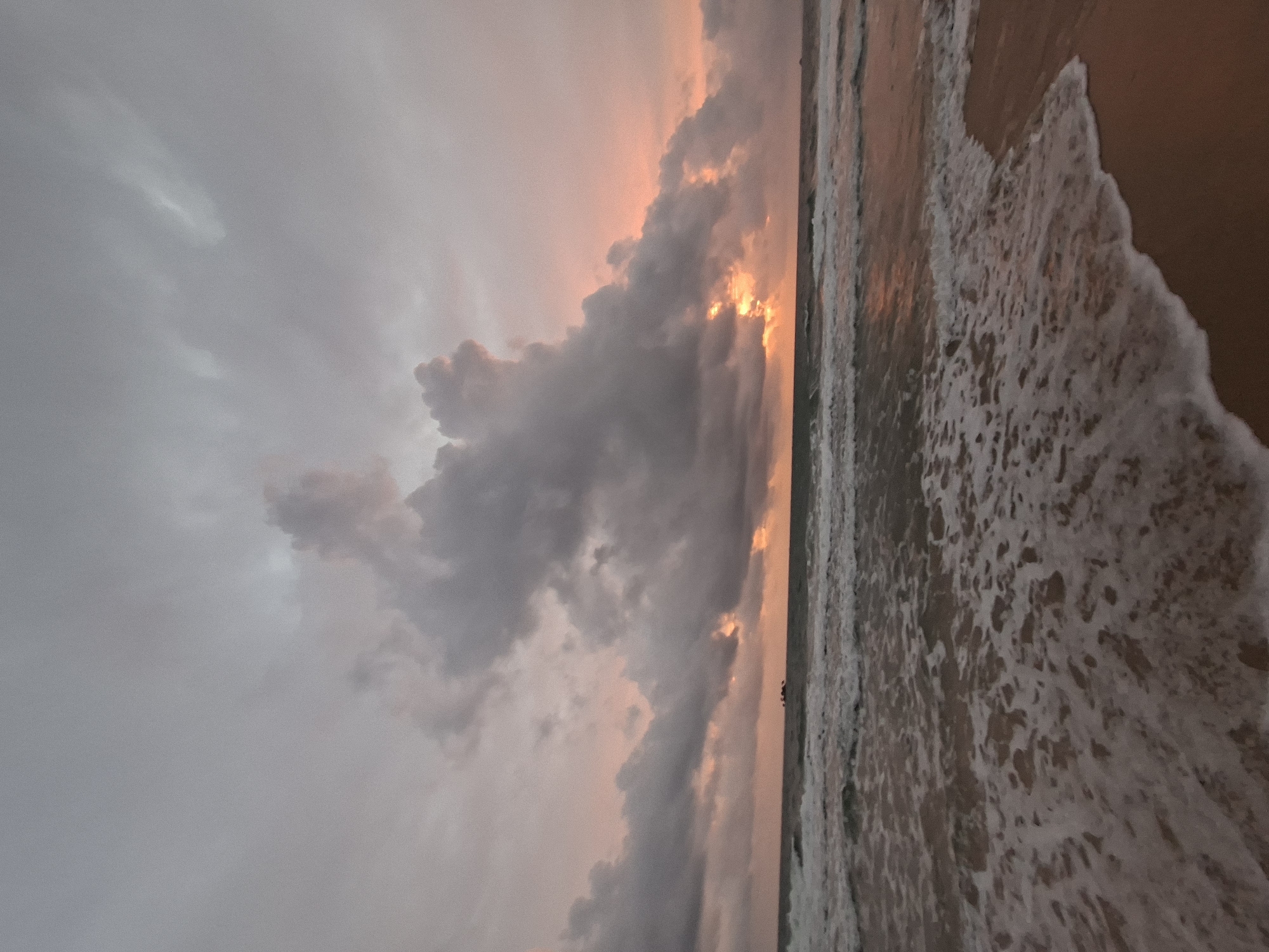 The image depicts a serene beach scene at sunset in Phú Quốc, Vietnam. The sky is filled with dramatic, overcast clouds, tinged with the warm hues of the setting sun, creating a diffused orange glow that pierces through gaps in the clouds. The ocean is calm with gentle waves lapping at the shore, creating foamy patterns on the wet sand. A faint silhouette of a small boat can be seen on the horizon. The overall atmosphere is tranquil and moody, characteristic of a tropical twilight.