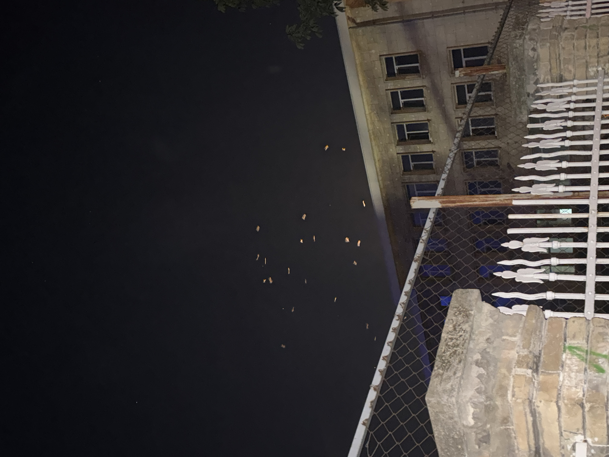An outdoor, nighttime, medium shot captures a dark sky above a building and fence. The sky is largely black, with a few small, faint lights that could be stars or distant aircraft. Scattered throughout the sky are numerous small, rectangular, illuminated objects, appearing to be floating or drifting. These objects are a pale orange or yellow color and vary in size and orientation. Below the sky is a light-colored, multi-story building with several windows on each floor. The building's facade appears to be made of stone or concrete blocks. A chain-link fence runs horizontally across the lower half of the image, with a weathered brick pillar on the left. The fence has metal posts, some of which appear rusted. Beyond the chain-link fence, there is a decorative white metal fence with pointed finials. The overall scene suggests a nighttime event involving floating lights, possibly lanterns, over an urban or suburban setting with a building and fencing in the foreground.