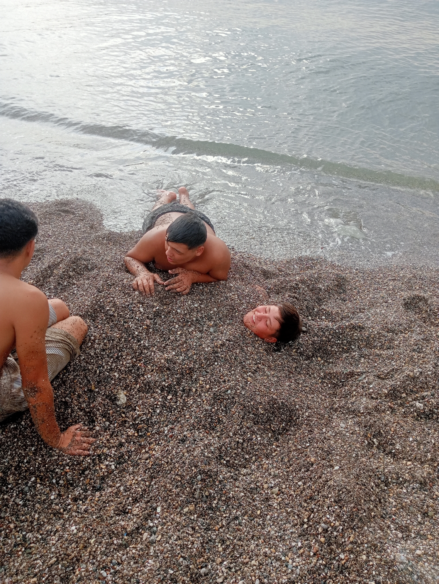 In Fethiye, Turkey, two young males are on a pebble beach. One individual is lying prone on the beach, with his body partially submerged in the pebbles, only his head and shoulders visible. He is smiling, with his eyes closed and his head tilted back. The other young male is seated on the beach, facing away from the viewer and toward the individual in the pebbles. He is wearing patterned shorts and his left arm is resting on the pebbles. The sea is visible in the background, with small waves lapping at the shore. The overall impression is one of a casual, playful scene on a sunny day at the beach.