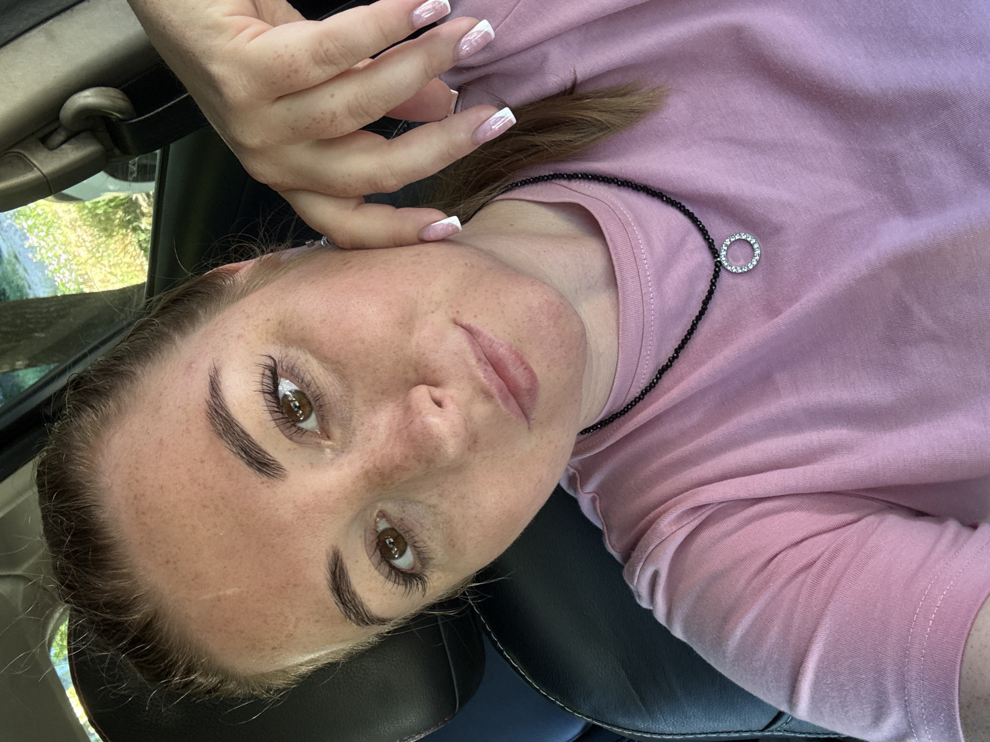 A medium shot photograph shows a woman with freckles, light brown hair pulled back, and brown eyes. She is wearing a pink t-shirt and a beaded necklace with a circular pendant. Her right hand is resting on her cheek, and her nails are painted with a French manicure. She appears to be in the driver's seat of a car, as a seatbelt buckle is visible behind her. The background suggests an outdoor environment, possibly with trees and foliage. The scene is well-lit, indicating it is daytime. The camera is positioned at eye level with the woman, looking slightly upwards.