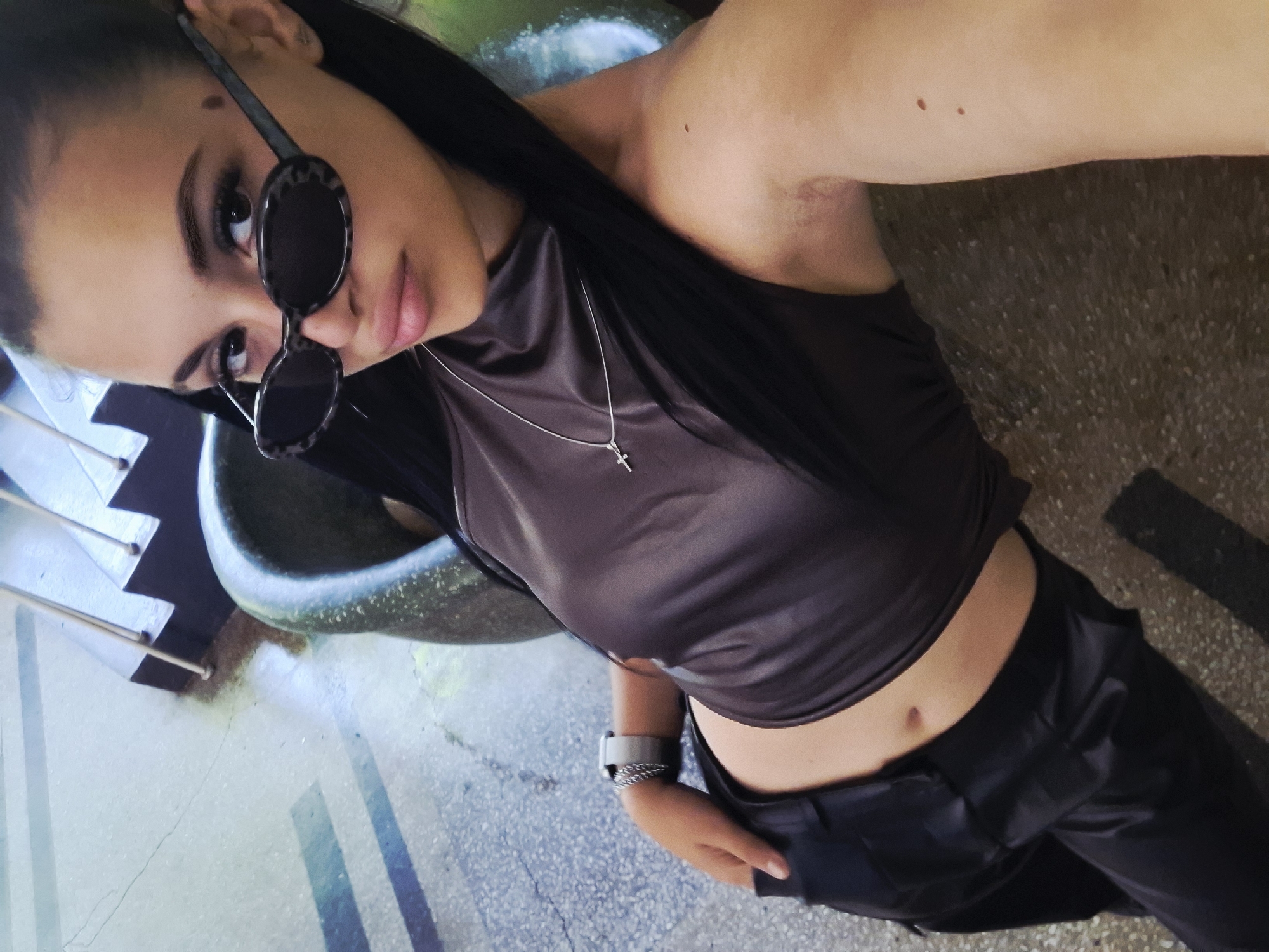 A young woman is captured in a high-angle, tilted portrait, looking upwards towards the camera. She appears to be in her late teens or early twenties, with long, dark hair pulled back. She wears dark, oval-shaped sunglasses with patterned frames, peering over them. Her attire consists of a dark, possibly satin or leather-look, sleeveless crop top that reveals her midriff, paired with matching dark trousers. A silver-colored chain necklace with a small cross pendant hangs around her neck. On her left wrist, she wears a light-colored watch and a silver-toned braided bracelet.
Notable facial features include defined eyebrows, dark eyes, and a small mole above her left eyebrow. Another mole is visible on her upper right arm. The setting appears to be outdoors in a brightly lit environment, suggesting daytime. The background features hints of concrete or paved ground with some abstract greenish and bluish elements, possibly part of urban infrastructure or public art. There are no visible texts in the image. The woman is posing for the photograph, her expression neutral. The location context for this image is Mezdra, Bulgaria.