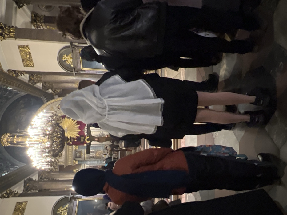 The image displays the interior of Saint Isaac's Cathedral in Saint Petersburg, Russia. The perspective is from behind several individuals standing on a tiled floor.
In the foreground-center, a person wears a black dress or skirt and a translucent white hooded poncho. Their bare lower legs are visible, ending in dark low-cut shoes. To the left, another individual, seen from the back, wears an orange-brown jacket, dark trousers, and a dark blue scarf draped over their head and left shoulder. They carry a patterned bag in their left hand and wear dark boots. To the right, a third person is seen from the back, wearing a dark leather jacket with a light-colored collar, dark trousers, and dark shoes. This individual has medium-length brown hair.
In the mid-ground, a large wooden crucifix with a golden halo behind the figure of Christ is visible, positioned near what appears to be an altar area, surrounded by other indistinct figures, some of whom are kneeling. Above, a prominent, multi-tiered, gilded chandelier with numerous lights hangs from the high, ornate ceiling. The architectural environment features massive polished columns with gilded Corinthian capitals, decorative arches, and religious artwork on the walls and upper structures, illuminated by ambient artificial light.