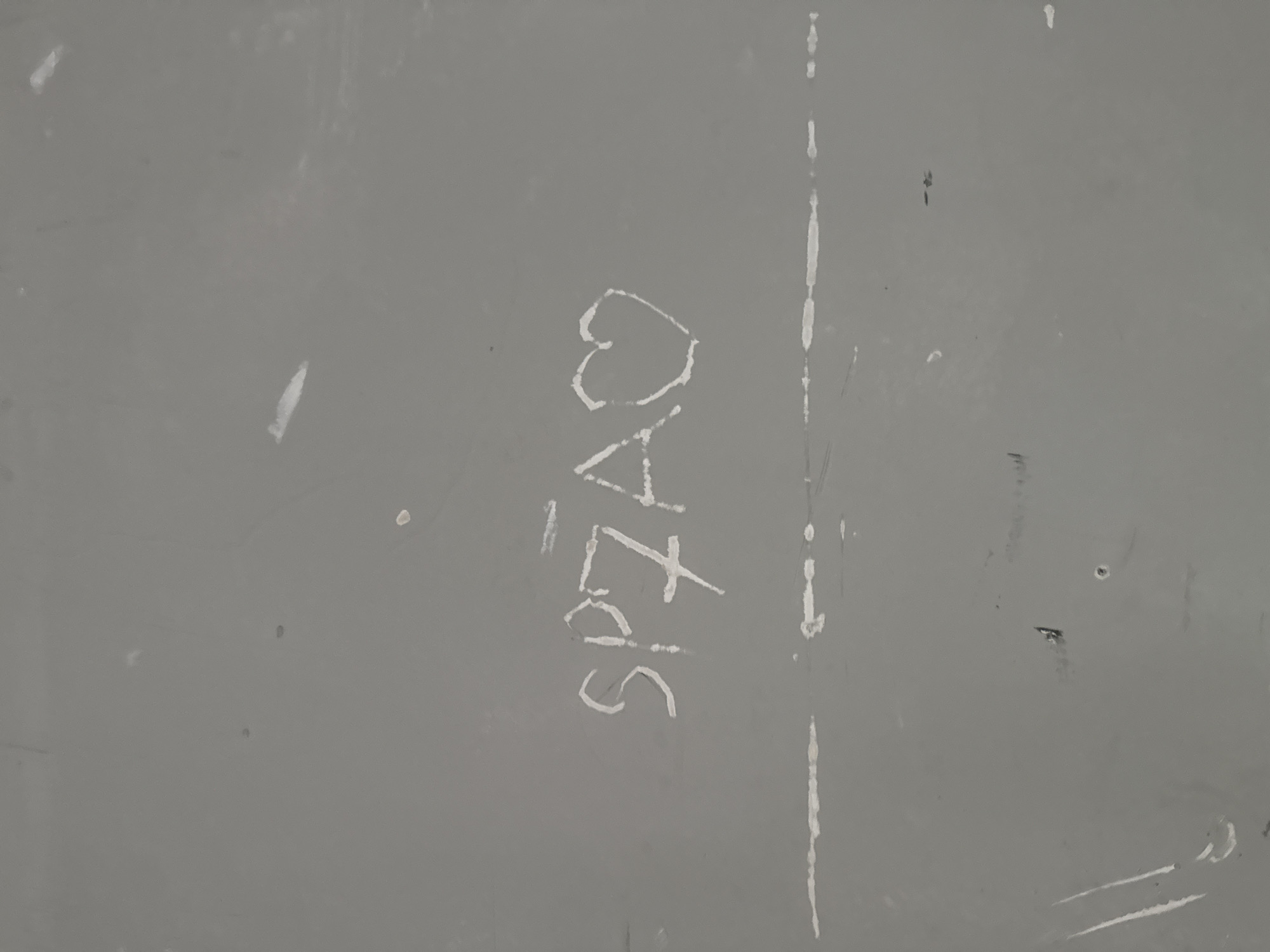 The image shows a section of a grey, textured wall. In the center of the frame, white chalk graffiti reads "SP7A" followed by a heart symbol. The graffiti is slightly smudged. Below the graffiti, a faint horizontal line, possibly another marking or scratch, stretches across the wall. The environment appears to be outdoors, given the texture and markings on the wall, but no specific urban features are visible to ascertain the exact location within Tbilisi, Georgia.