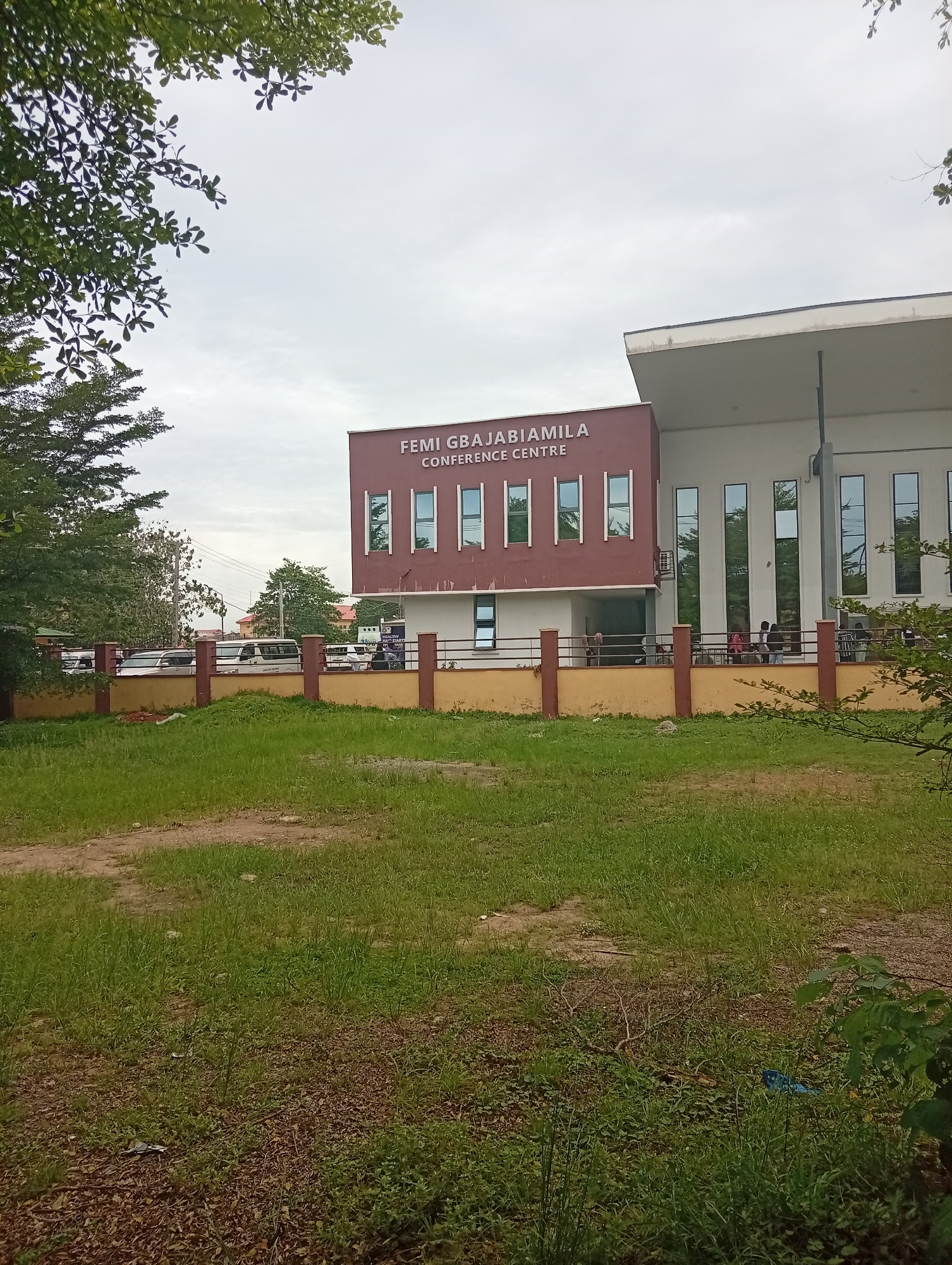 The image depicts the Femi Gbajabiamila Conference Centre in Ijanikin, Nigeria. The conference centre is a two-story building with a maroon section and a white section. The building has several vertical windows. A brown and yellow fence runs along the front of the building. People are visible standing near the entrance. A grassy area is in the foreground, with trees partially obscuring the view on the left and right sides of the frame. Several vehicles are parked behind the fence. The sky is overcast.