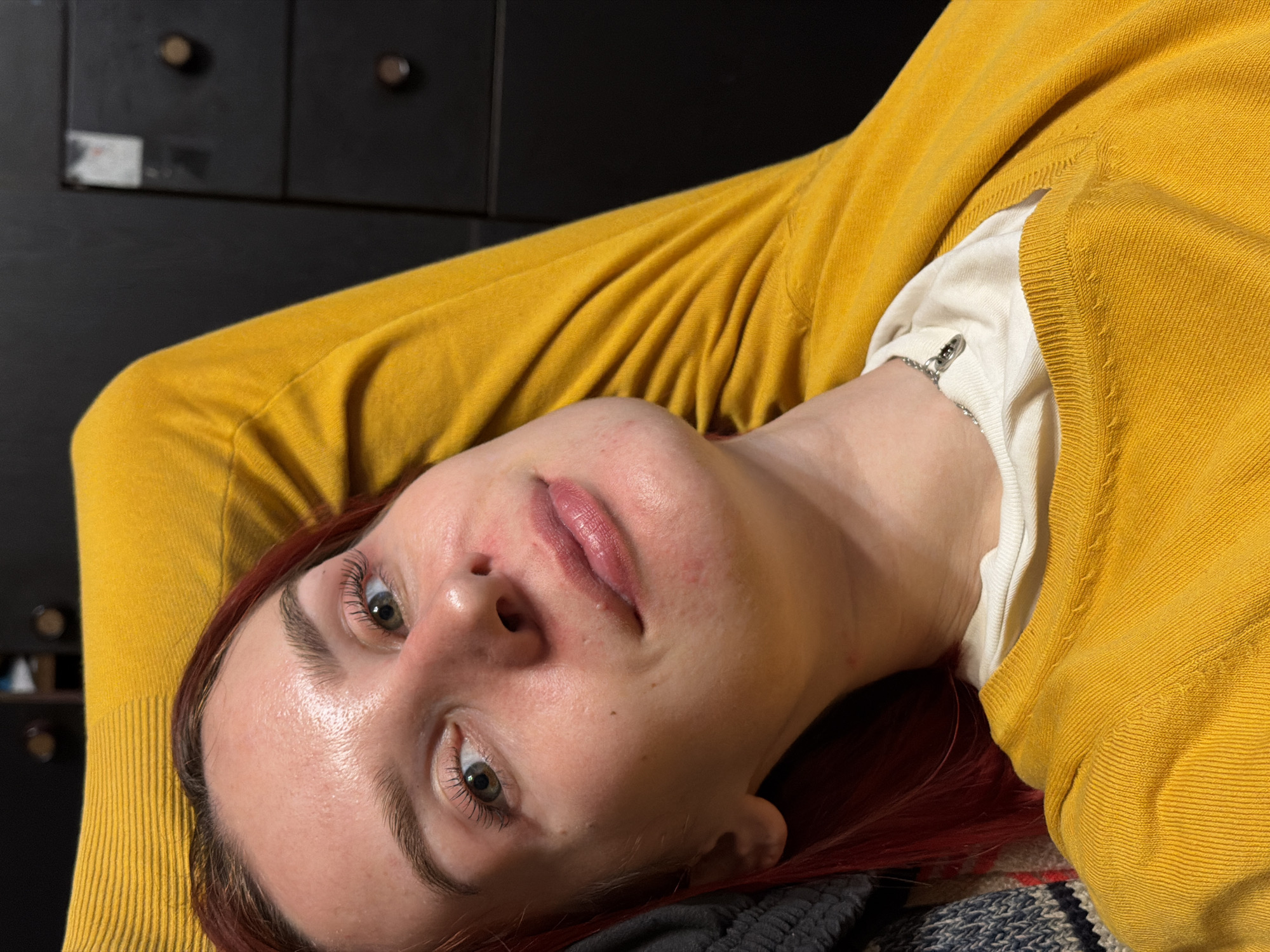 The image presents a close-up, overhead portrait of a person, likely a woman, with reddish-brown hair. She is depicted lying down or reclined, with her head tilted to the side, wearing a mustard yellow long-sleeved sweater over a white undershirt. A silver chain is visible on her left wrist, peeking out from beneath the white sleeve.
Her face is the primary focus, revealing fair skin that appears somewhat shiny or dewy. She has defined eyebrows and dark eyelashes, and her eyes, which are open, gaze directly towards the viewer. Her lips are slightly parted, showing a natural pink color, and there are a few small red marks or blemishes visible on her nose and chin.
The background is dark and largely out of focus, suggesting an indoor setting. A dark piece of furniture, possibly a chest of drawers, with two small, light-colored round knobs, is visible behind her upper body. To the left of these knobs, a small, blurry, light-colored sticker or label is present on the dark surface, but no text is discernible. In the bottom right corner of the frame, a segment of patterned fabric, potentially a blanket or another piece of clothing with blue, white, and red stripes, is visible. The scene conveys a relaxed and calm atmosphere, appearing to be a casual portrait or a moment of rest in Заболотово, Russia.