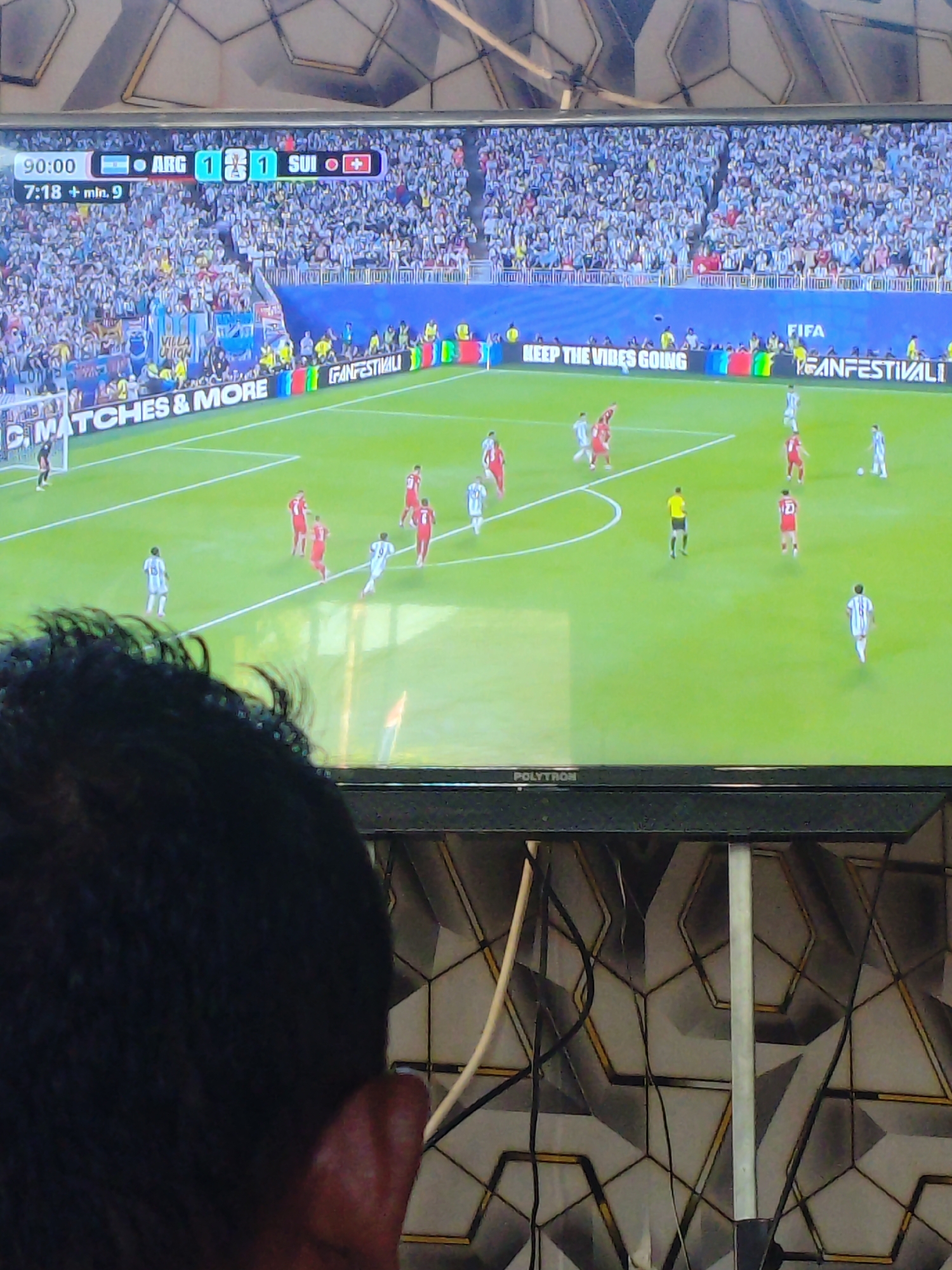 The image shows a close-up view of a television screen displaying a soccer match. The score is 1-1 between ARG and SUI. The time is 90:00, with 7:18 + min. 9 indicating stoppage time. The game is being played in a stadium with a large crowd in the stands, most of whom are wearing light blue shirts, likely supporting the Argentinian team. The field is green with white markings. There are players in red and light blue uniforms on the field, actively engaged in the game. Advertisements are visible along the edge of the field, including "MATCHES & MORE" and "KEEP THE VIBES GOING". The lower part of the image shows the top of a television set with the brand name "POLYTRON". The background behind the television is a wall with a geometric pattern. The image is captured from behind and slightly to the side of a person, whose dark hair and ear are visible in the foreground, suggesting the television is being watched in a room. The question of location in Parangsembilan, Indonesia, cannot be determined from the visual information provided in the image.