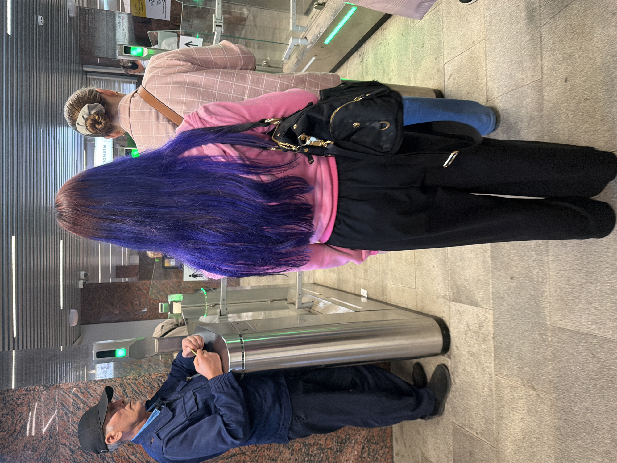 In the metro, a man in a dark blue uniform and a black cap stands next to a turnstile, holding a yellow ticket. The turnstile has a green light indicating it is operational. To the right of the man is a woman with long, bright purple hair, wearing a pink hoodie and black pants, carrying a black handbag. Further to the right, another woman with her hair in a bun wears a pink plaid shirt and blue jeans, with a tan strap from her bag visible. The scene appears to be a metro station entrance with granite-tiled flooring and marble-patterned walls.