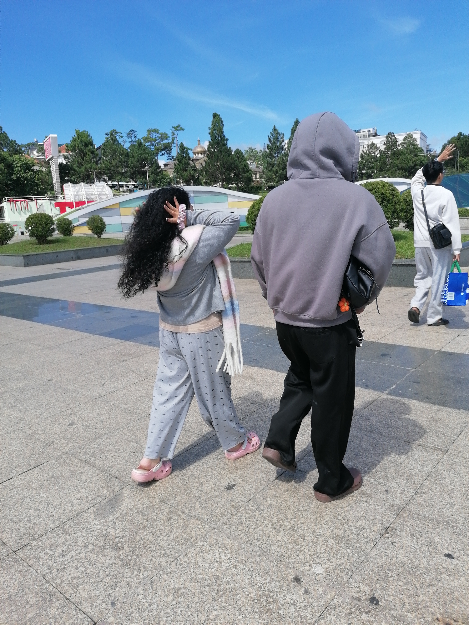In Da Lat, Vietnam, on a paved plaza under a clear blue sky, three individuals walk away from the camera. The person in the foreground, from the back, has long curly dark hair and wears a gray hooded sweatshirt over a pale undershirt, black pants, and dark clogs. A black bag is slung over their shoulder, and an orange keychain dangles from the bag strap. Beside them, a person with dark curly hair pulled back with a scrunchie wears a gray long-sleeved shirt, a colorful striped scarf, patterned light gray pajama pants, and pink clogs. They are touching their hair with one hand. In the background, another person in a white sweatshirt and light gray pants, carrying a blue shopping bag, walks away with their right arm raised. In the distance, lush green trees and modern buildings form the city skyline.