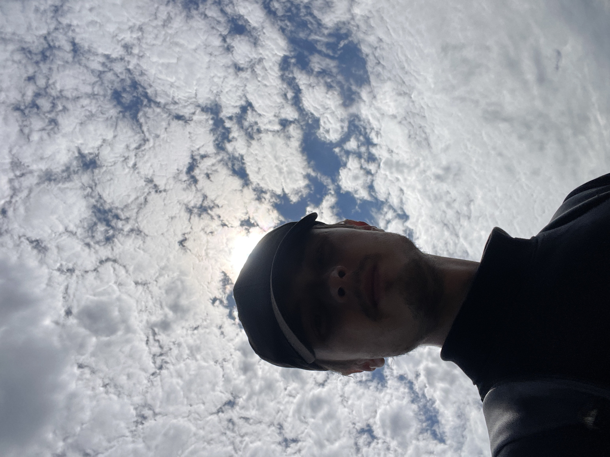 A low-angle, head-and-shoulders selfie is captured against a cloudy sky with the sun partially obscured. The subject, a young person wearing a black baseball cap and a black turtleneck shirt, is looking directly at the camera. Their face is in shadow, with only the lower part of their nose and a faint mustache and goatee visible. The background is dominated by a dense layer of cumulus clouds, interspersed with patches of blue sky. The image appears to be taken outdoors, possibly in Diepenbeek, Belgium, given the context provided. There are no other individuals or objects of significance visible in the frame.