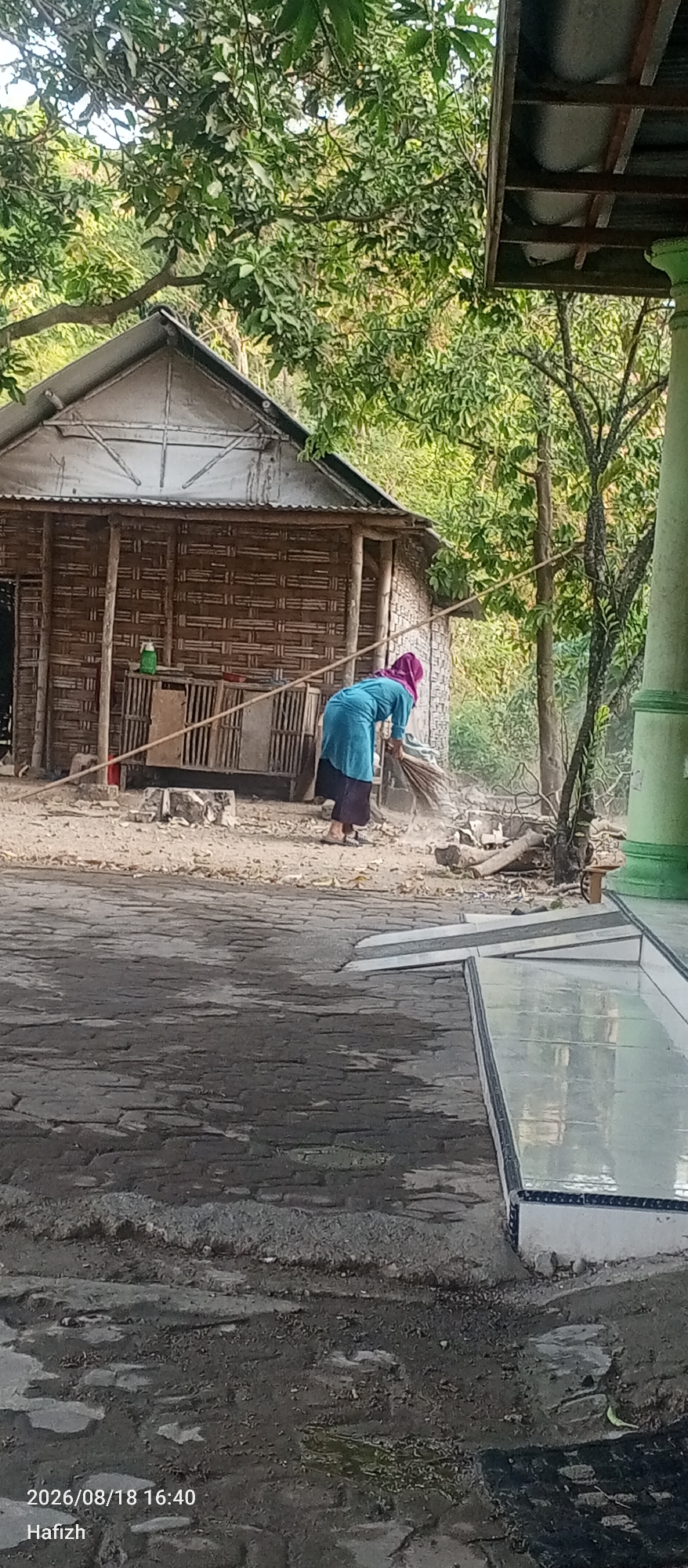 In Lebanisuko, Indonesia, a woman wearing a purple headscarf and a turquoise abaya is sweeping the dirt ground in front of a bamboo-walled house. The house has a corrugated metal roof and a wooden frame. The ground is covered in dirt, leaves, and small stones. A green bottle is visible on a shelf inside the house. To the right of the house, a green pillar stands near a tree with green leaves. The foreground shows a paved area with a reflective surface.