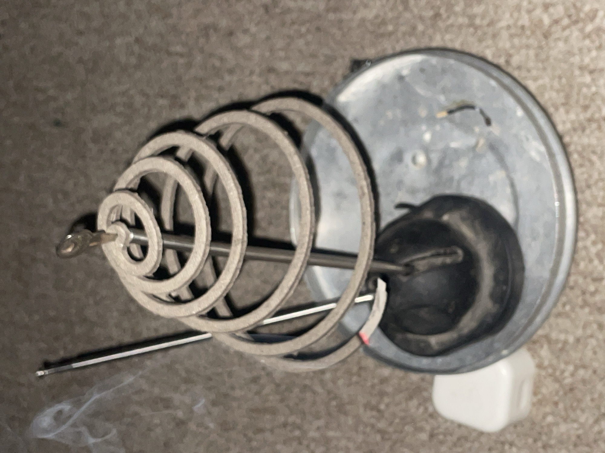 The image shows a close-up of a mosquito coil burning in a metal holder. The coil is spiraled and appears to be made of a light-colored material. A thin stick, likely an incense stick, is placed next to the coil and is producing a plume of white smoke. The holder is round and made of metal, with some ash and burnt residue on its surface. To the left of the holder, there is a small white square object. The background appears to be a carpeted surface. The overall scene suggests a quiet, indoor setting where the mosquito coil is being used for pest control or ambiance. There are no people or animals visible in the image. The image does not provide any information about the location in Jalingo, Nigeria.