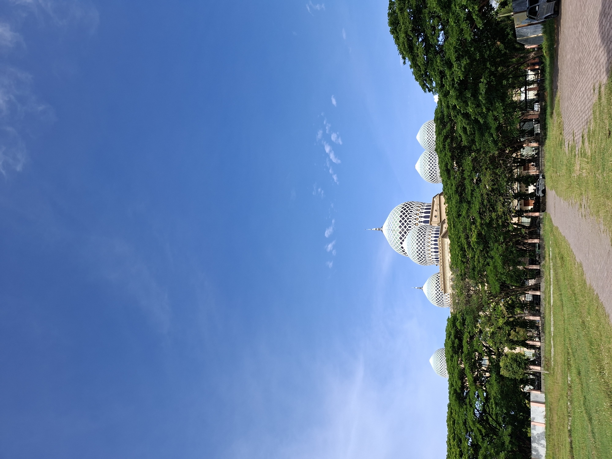 The image shows Baiturrahman Mosque, viewed from ground level in Lhokseumawe, Indonesia. The sky is clear blue with wisps of white clouds. Several domes, distinguished by their white, latticed design, are visible above a line of trees. Adjacent to the trees is a paved walkway and a grassy area. A portion of a building is visible behind the trees. A car is parked on the walkway.