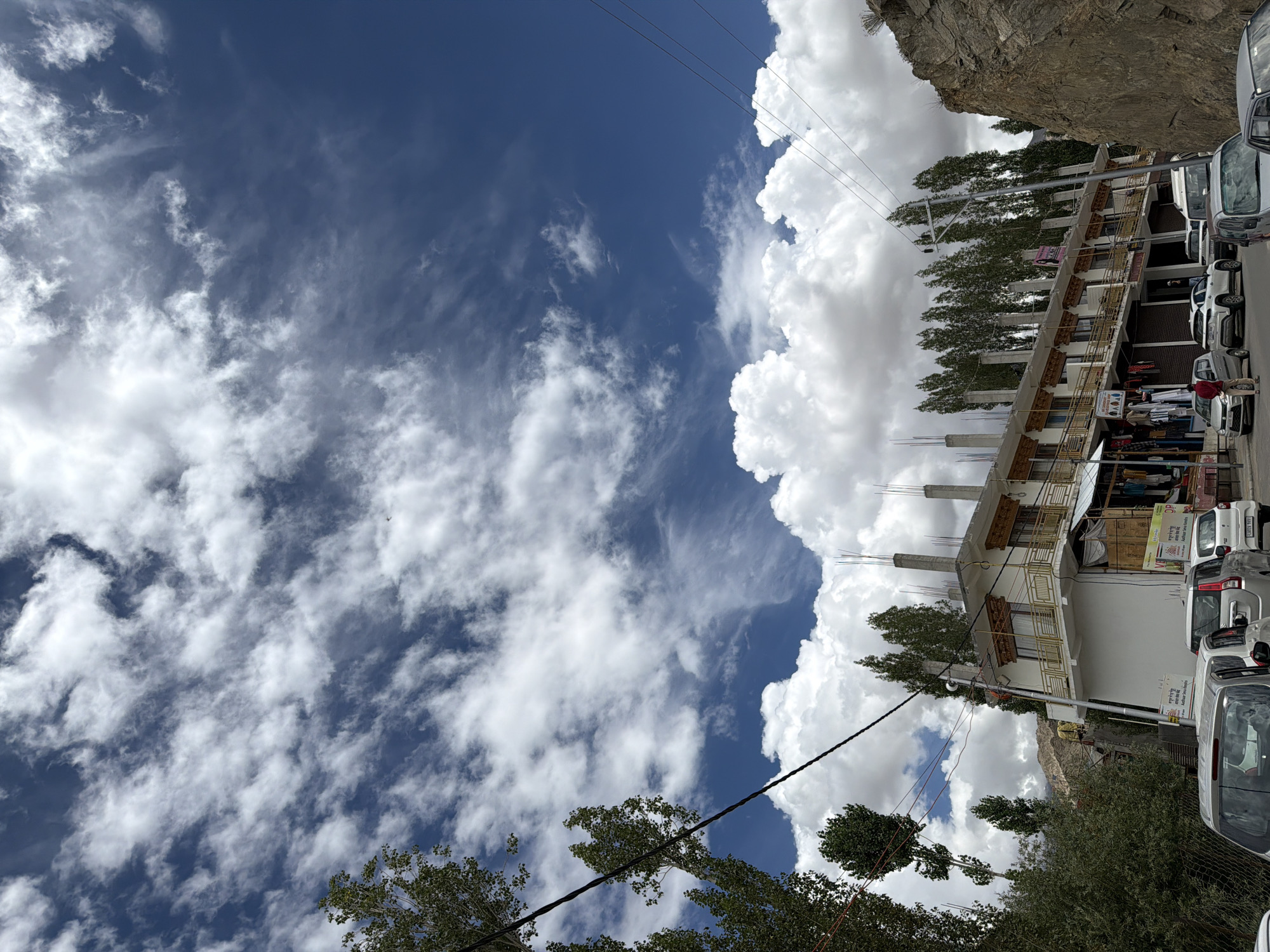 The image depicts a street scene in Leh, India, characterized by a blue sky filled with white, wispy clouds. On the left side, tall trees with green foliage are visible. A multi-story building with white walls and a tiled roof, featuring balconies with ornate railings, lines the street. The building appears to be a commercial establishment, with storefronts displaying clothing and other goods on the ground floor. Several cars are parked along the street, and a few individuals can be seen. In the background, a rocky hillside is present, adding to the mountainous landscape typical of Leh. Power lines crisscross the scene, and some construction work is evident on the building's upper levels, with concrete pillars protruding. The overall impression is of a vibrant, busy street in a town set against a dramatic natural backdrop.