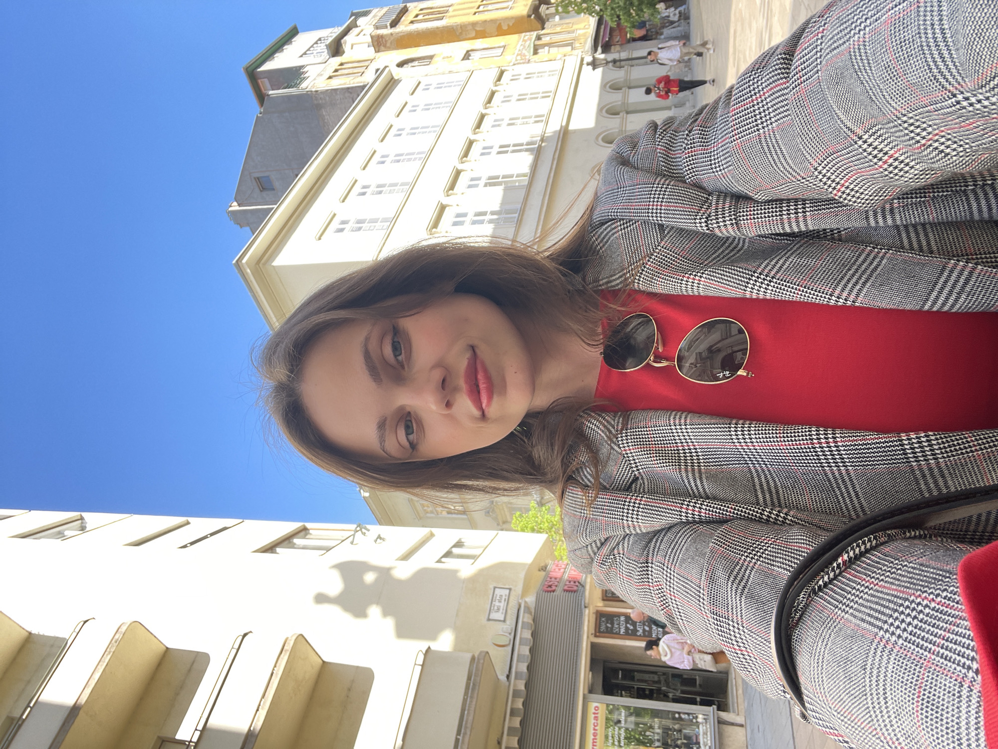 A young woman is standing outside, taking a selfie. She wears a red shirt with a patterned gray and red blazer. Sunglasses are clipped to the shirt. In the background is a city street with buildings. One business is a market called "Mercato". This is in Budapest, Hungary. There are other people walking in the background.