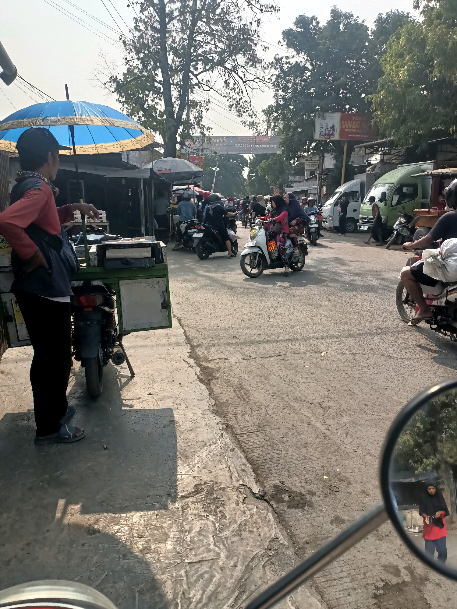 A sunny day scene unfolds in Ujungrusi, Indonesia, where a street is lined with small businesses and bustling with motorcycle traffic. In the foreground, a person stands by a parked motorcycle with a green storage box attached, facing away from the camera. The person wears a red shirt and black pants, with a blue bag slung over their shoulder. A large blue and yellow umbrella is set up nearby, casting a shadow. The street is paved with asphalt and shows signs of wear. In the mid-ground, several motorcycles, some with riders, navigate the road. One white scooter carries two women wearing hijabs. Further down the street, a green truck and a white van are parked. Overhead, power lines crisscross the sky, and trees with dense foliage line the roadside. A billboard with Indonesian text and images of people is visible in the background. The overall impression is one of a lively, everyday street scene in a tropical urban environment.