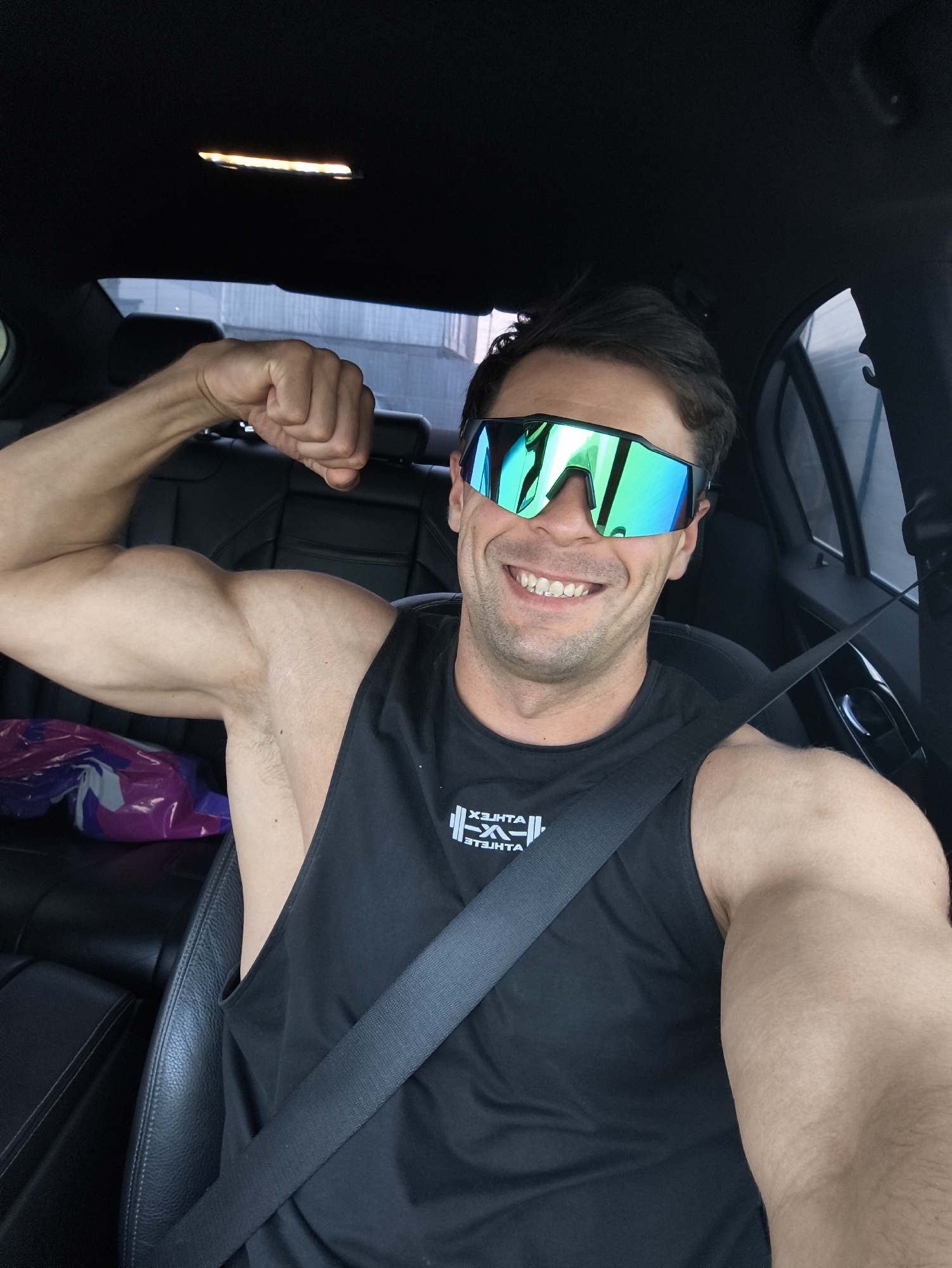 A male individual is depicted seated in the driver's seat of a vehicle with a dark interior, located in Molokovo, Russia. He wears large, reflective green-blue mirrored sunglasses and a black sleeveless tank top featuring a reversed white logo: "X3HTA" above a stylized barbell graphic and "ATHLETE". The man is smiling, exposing his teeth, and is secured by a dark seatbelt across his chest and lap. His right arm is flexed at the elbow, displaying a bicep and clenched fist. A black leather headrest is visible in the rear passenger seat, behind his right arm, along with a purple and grey plastic bag. An amber-illuminated overhead light fixture is visible on the black headliner. The exterior, visible through the vehicle's windows, appears to be a blurred urban or semi-urban environment with a light-colored building structure, indicating an outdoor, daytime setting.