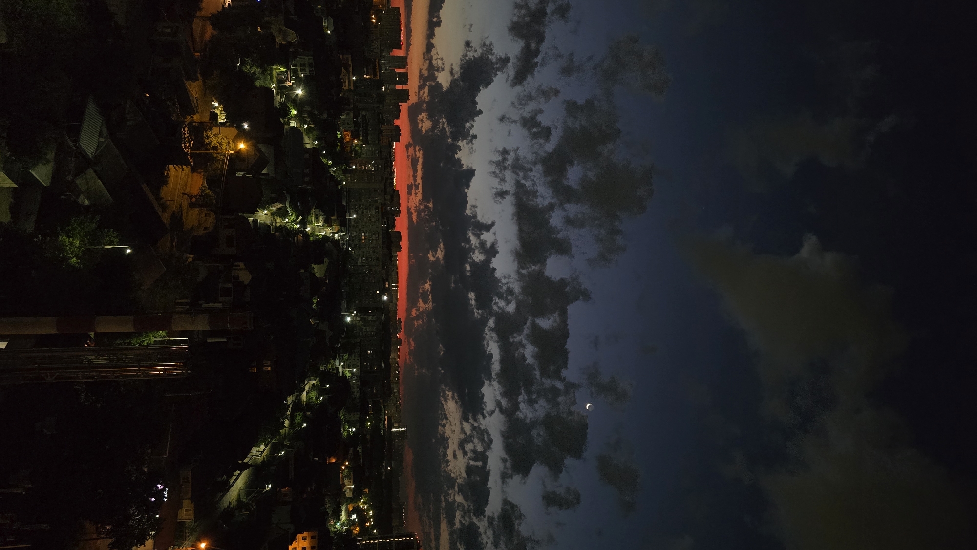 An aerial, high-angle, vertical shot captures a twilight cityscape with a crimson horizon. Dark, cumulus clouds are scattered across a deep blue sky, with a sliver of a crescent moon visible in the upper right quadrant. Below the sky, a dense collection of apartment buildings and smaller houses stretches across the frame, illuminated by the glow of streetlights and windows. In the foreground, a red and white striped smokestack stands tall, partially obscured by the dark shapes of trees. The overall impression is one of a city transitioning from day to night, with a mix of dense urban development and residential areas. This scene is consistent with a view from a high vantage point overlooking a district within the city of Krasnodar, Russia, such as a residential area near an industrial zone.