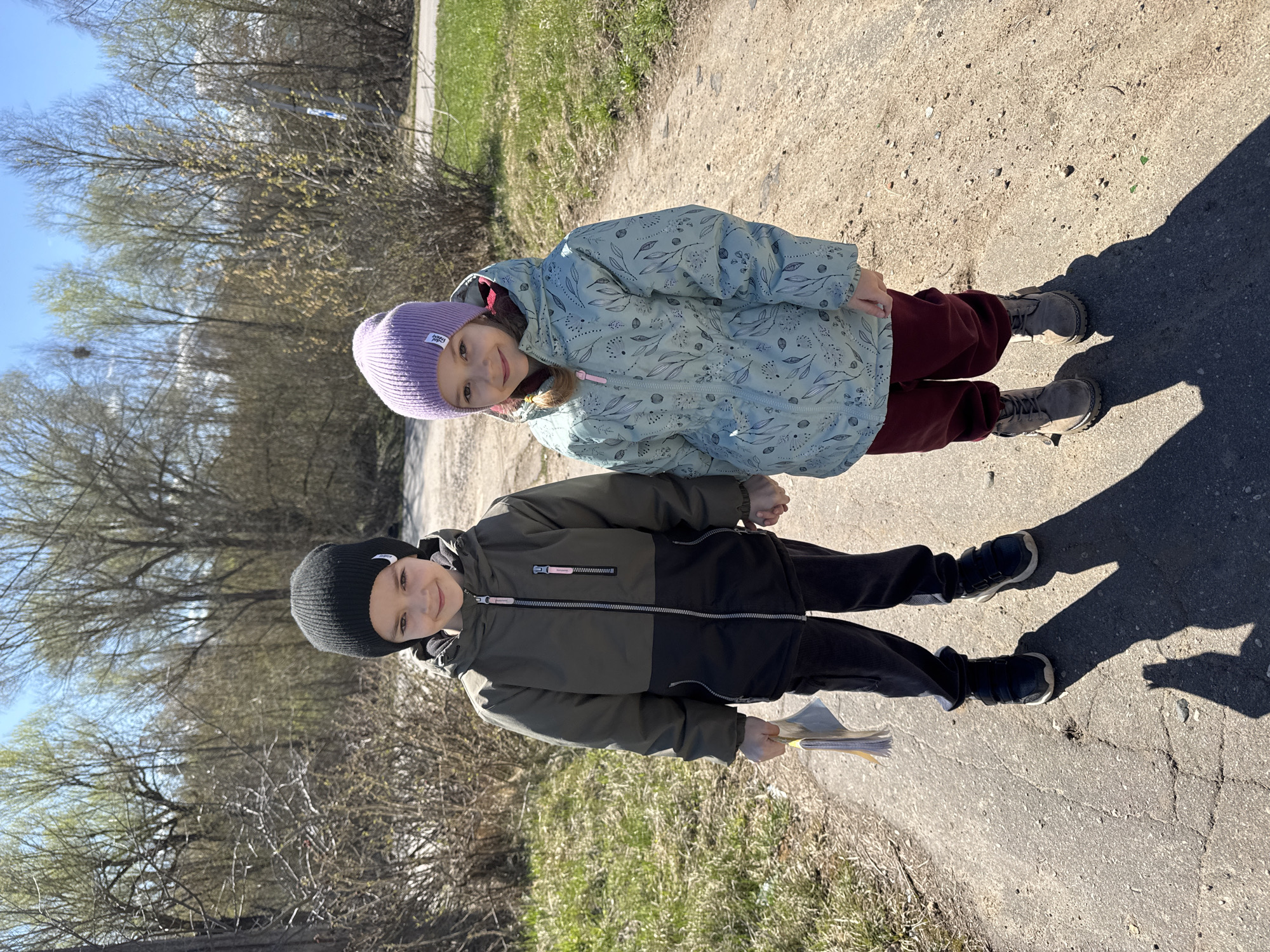 Two children, a girl and a boy, stand on a cracked concrete path in a sunny outdoor setting. The girl, positioned slightly behind the boy, wears a light blue patterned jacket, maroon pants, tan boots, and a purple beanie. The boy wears a two-tone green and black jacket, black pants, black sneakers with white soles, and a black beanie. He holds a stack of papers in his left hand. Both children are looking at the camera and smiling. The background consists of bare trees, grass, and a clear blue sky. The presence of bare trees and light clothing suggests a transition season like early spring. The location, Shevlyakovo, Russia, is not discernible from the image.