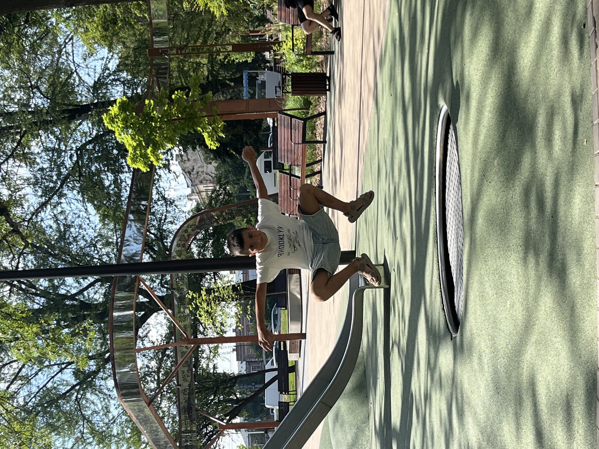 This is a vertical, medium shot image taken outdoors on a sunny day. The main subject is a young boy, likely a teenager, in mid-air, jumping or falling from a curved silver slide. He is wearing a white t-shirt with the text "BROOKLYN" printed on it, along with light blue shorts and beige sandals. His arms are outstretched, and his expression appears to be one of surprise or excitement.
The slide is part of a playground structure, with a green rubberized surface surrounding it. Shadows of trees are cast across the green surface, indicating a sunny afternoon. In the background, there are trees with lush green foliage, park benches, and a cylindrical trash bin. Some buildings are visible in the distance, suggesting an urban park setting. The overall scene conveys a sense of fun and activity. There are also reflective elements in the background, possibly part of the playground equipment, which distort the reflections of the trees and sky. The location is identified as Chernivtsi, Ukraine.
