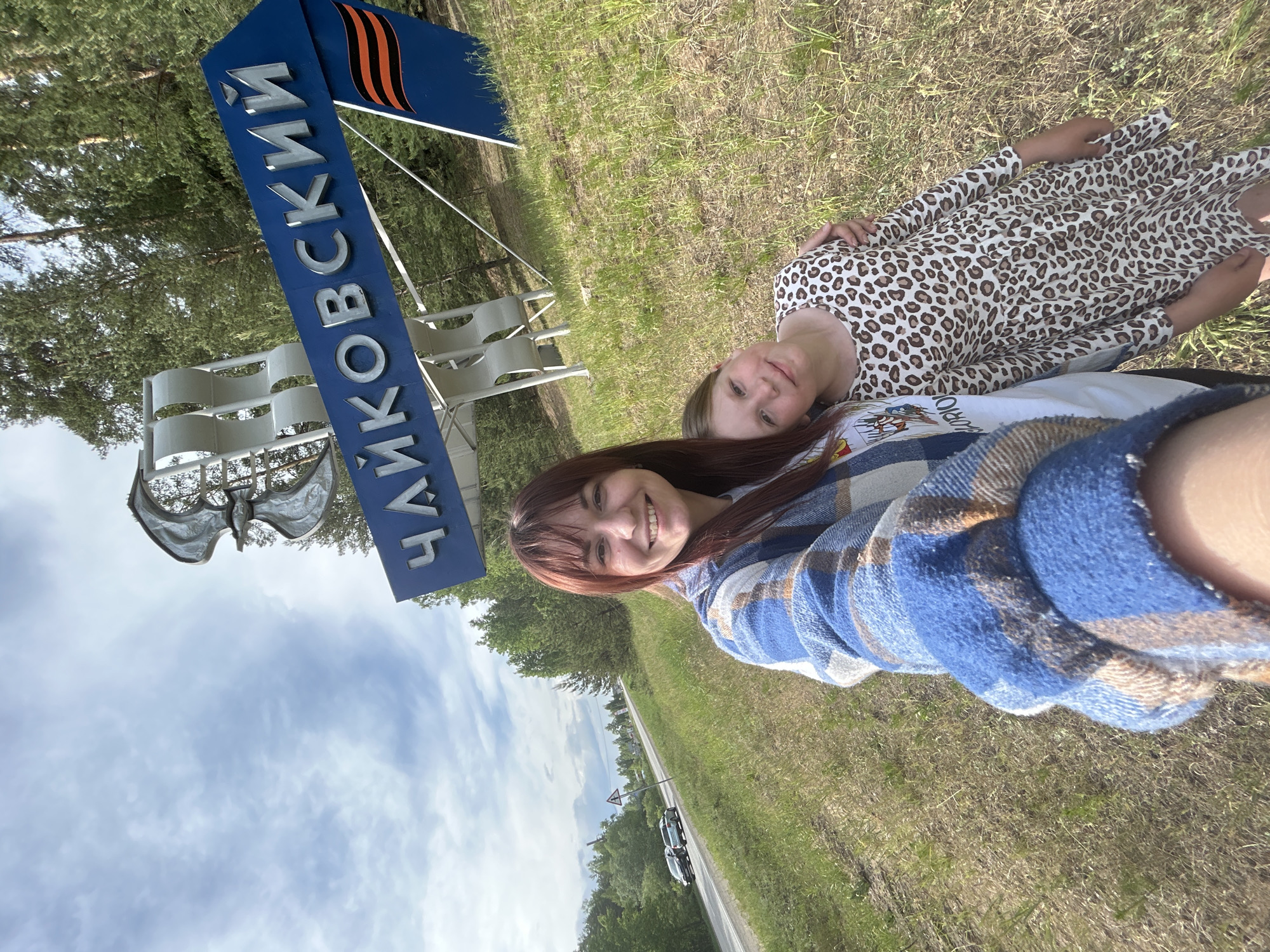 Two females are posing in a selfie in the city of Chaikovsky, Russia. One female, smiling, is closer to the camera and wearing a blue and brown plaid shirt. The other female, lying on the ground, is wearing a light-colored dress with brown spots. Behind them is a sign that reads "Чайковский" (Chaikovsky) in blue letters, topped with a depiction of a sailing ship and a stylized flag. The background includes a road with vehicles, a grassy area, trees, and a sky with clouds.