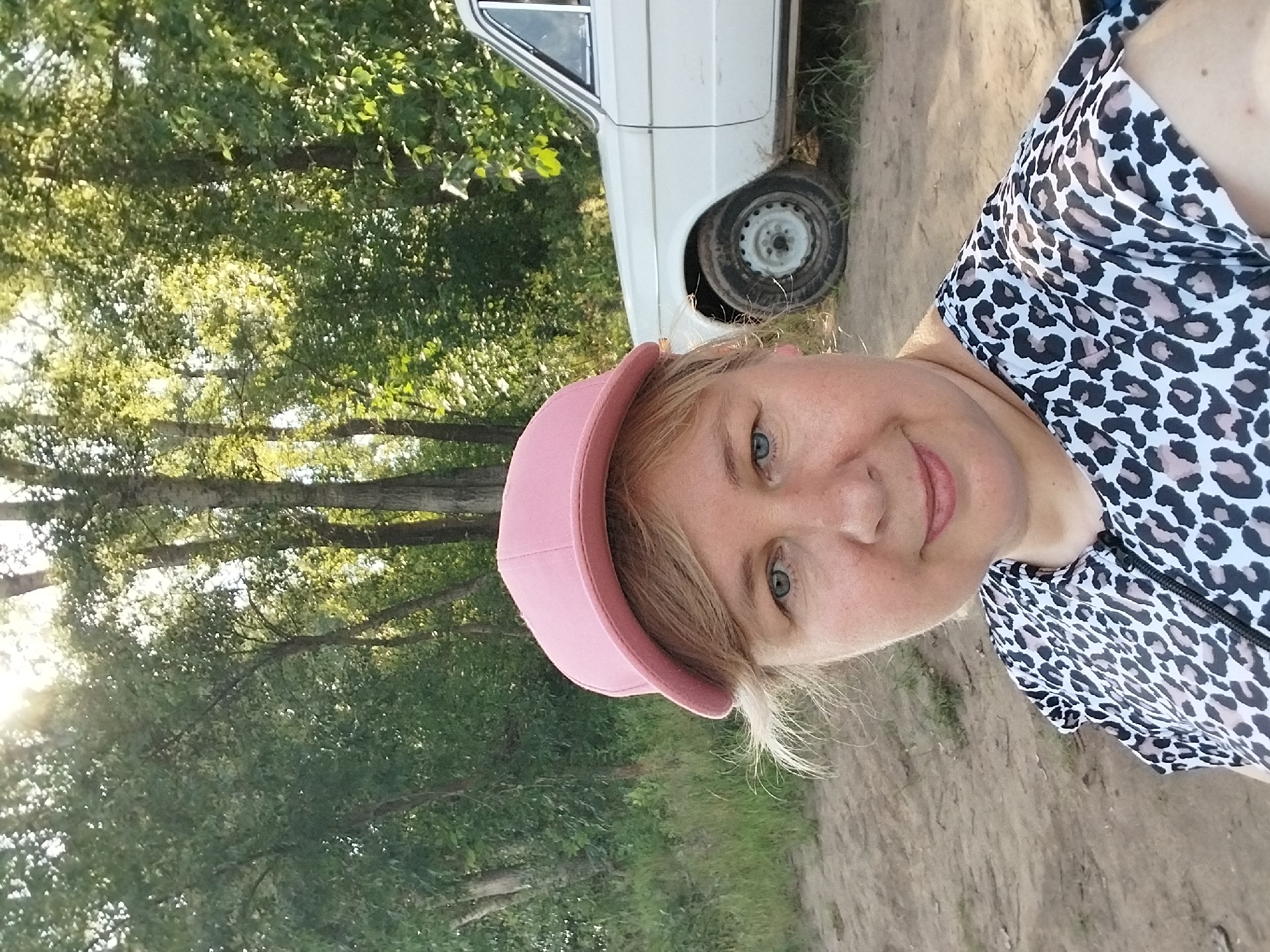 A woman wearing a pink baseball cap and a leopard-print top is standing in front of a white car. The car is an older model, possibly from the Soviet era, with a spare tire mounted on the side. The background consists of trees and foliage, suggesting an outdoor setting. The lighting indicates it is daytime, possibly late afternoon, with the sun filtering through the trees. The ground appears to be dirt or sand. The location is identified as Chkalov, Russia, though no specific city landmarks are visible. The woman is looking towards the camera, and her expression is neutral.